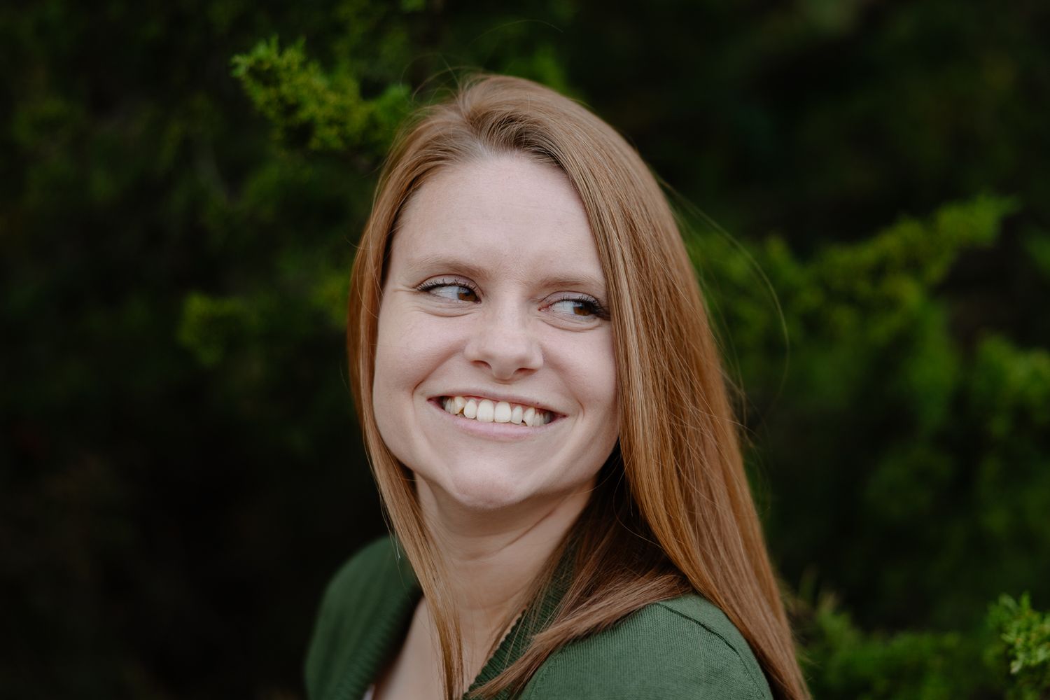 Portrait of someone in a green sweater smiling warmly against a natural green background.