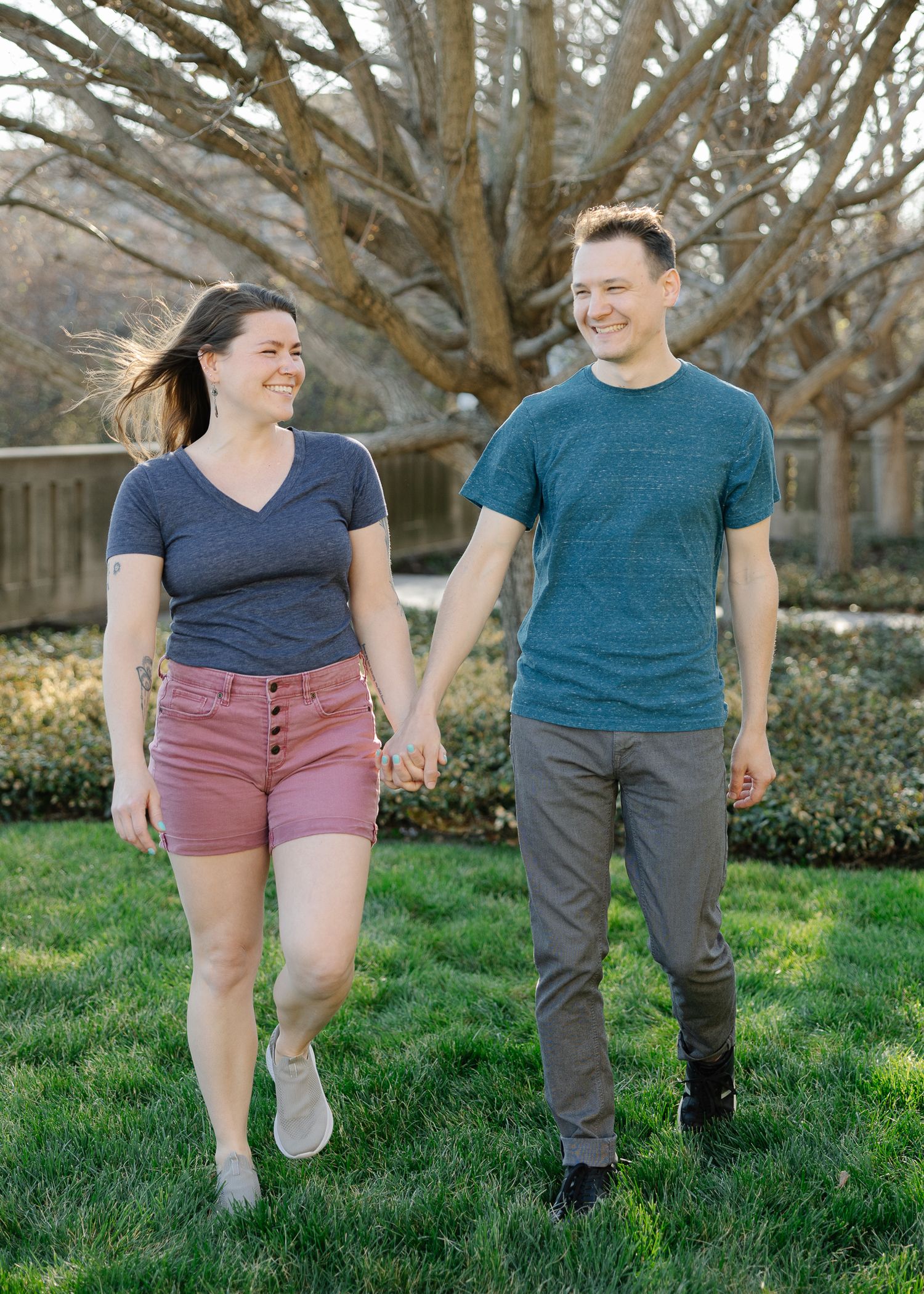 Couple holding hands and smiling while walking on green grass outdoors near bare trees on a sunny day.