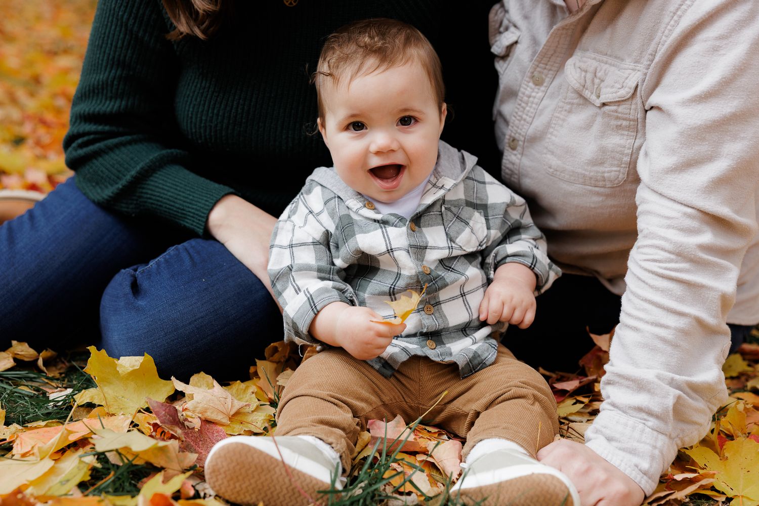 Happy baby in plaid shirt sitting on autumn leaves surrounded by family during fall outdoor photo session.