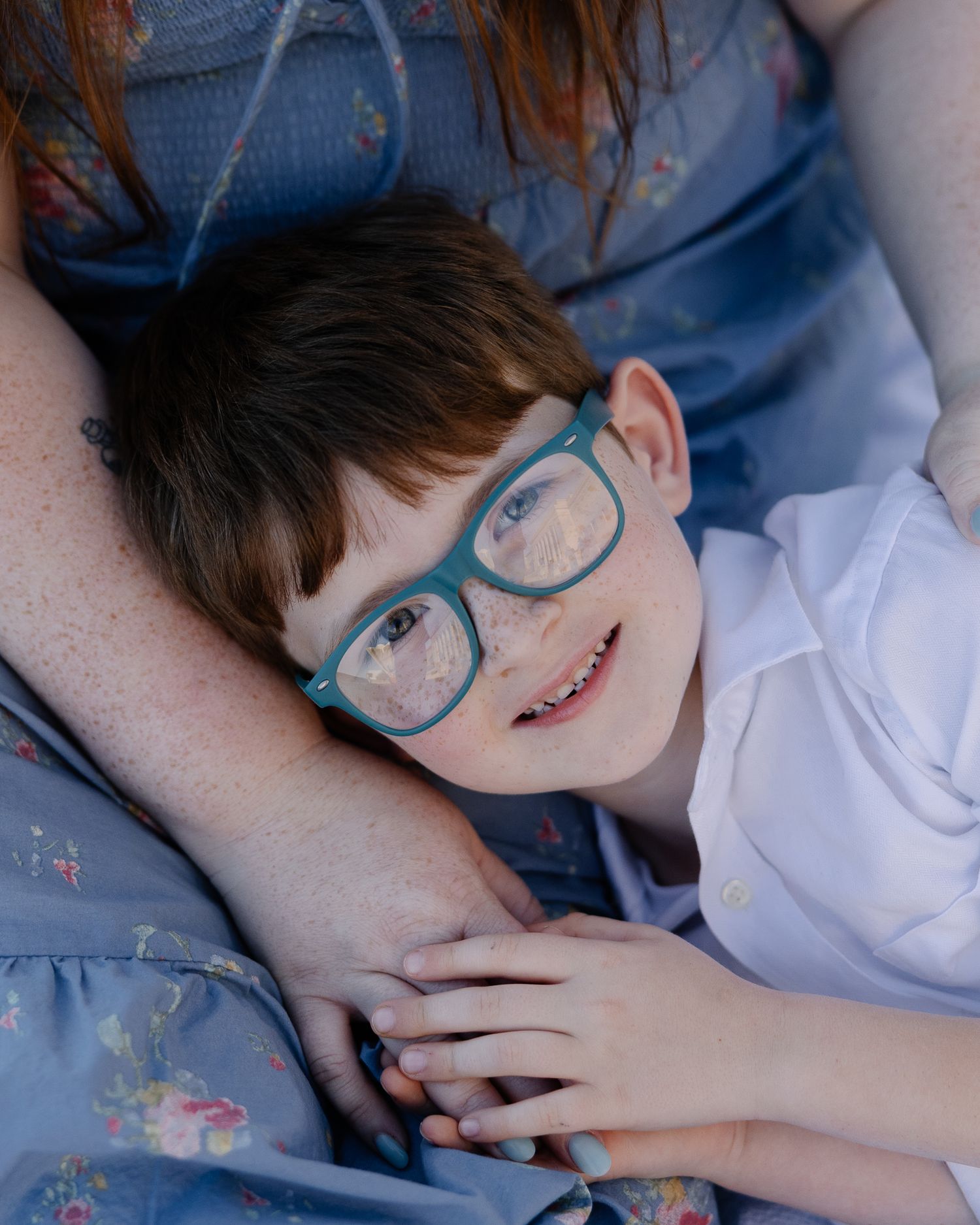 A smiling red-haired child wearing teal glasses rests their head on an adult's arm outdoors.
