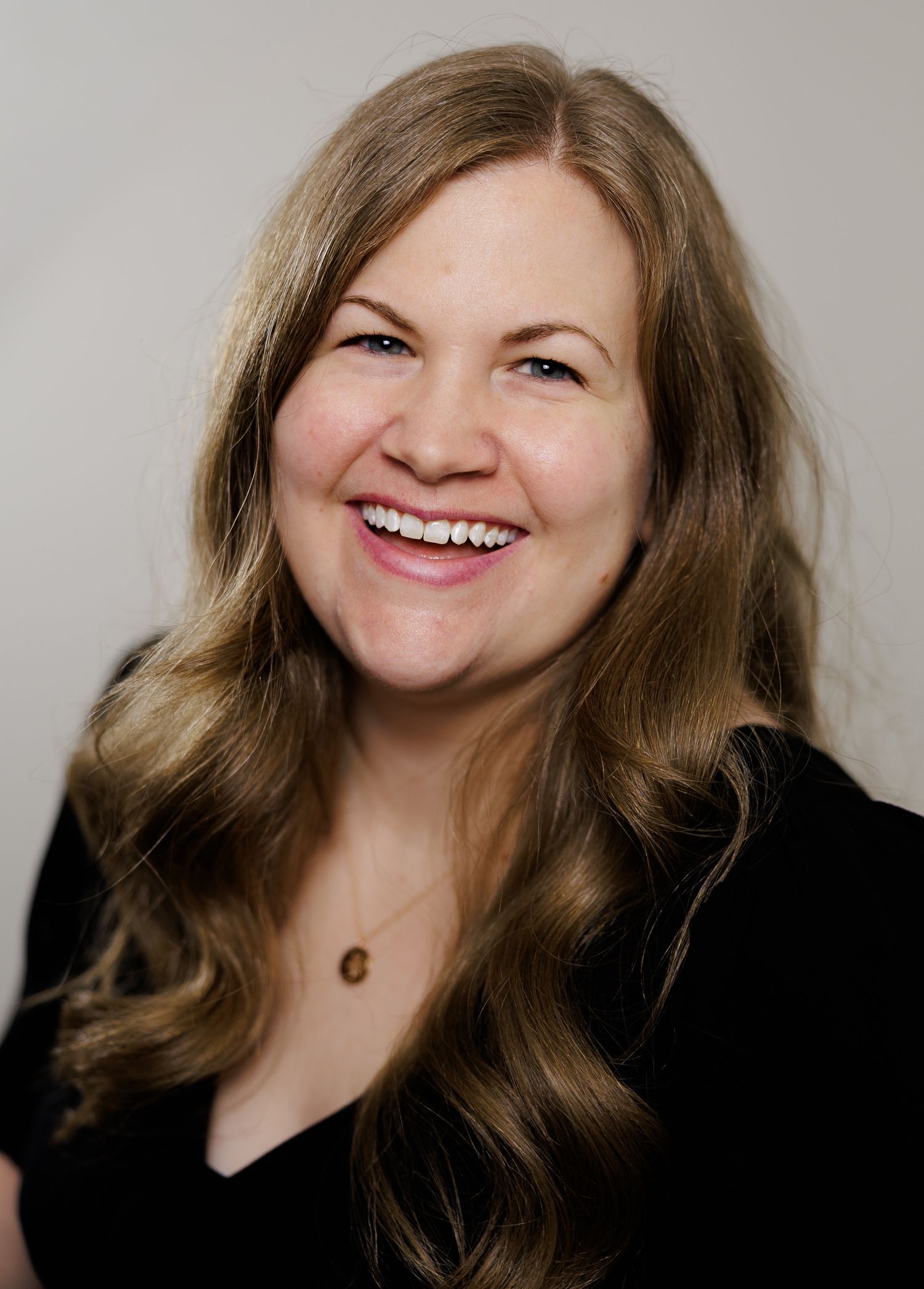 Smiling woman with long blonde hair wearing a black top and gold necklace against a neutral background.
