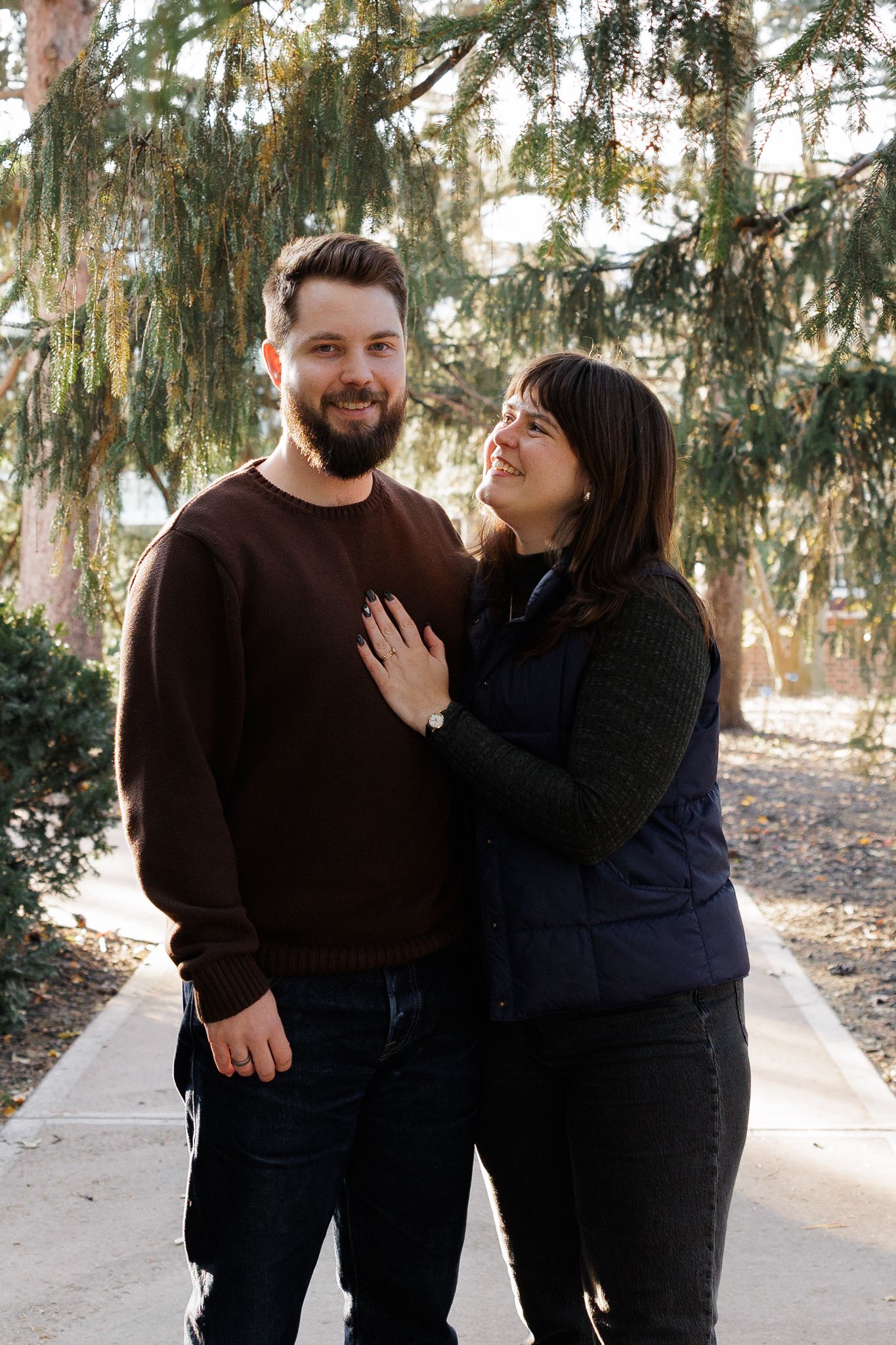 A smiling couple poses outdoors under evergreen trees, the woman resting her hand on the man's chest.