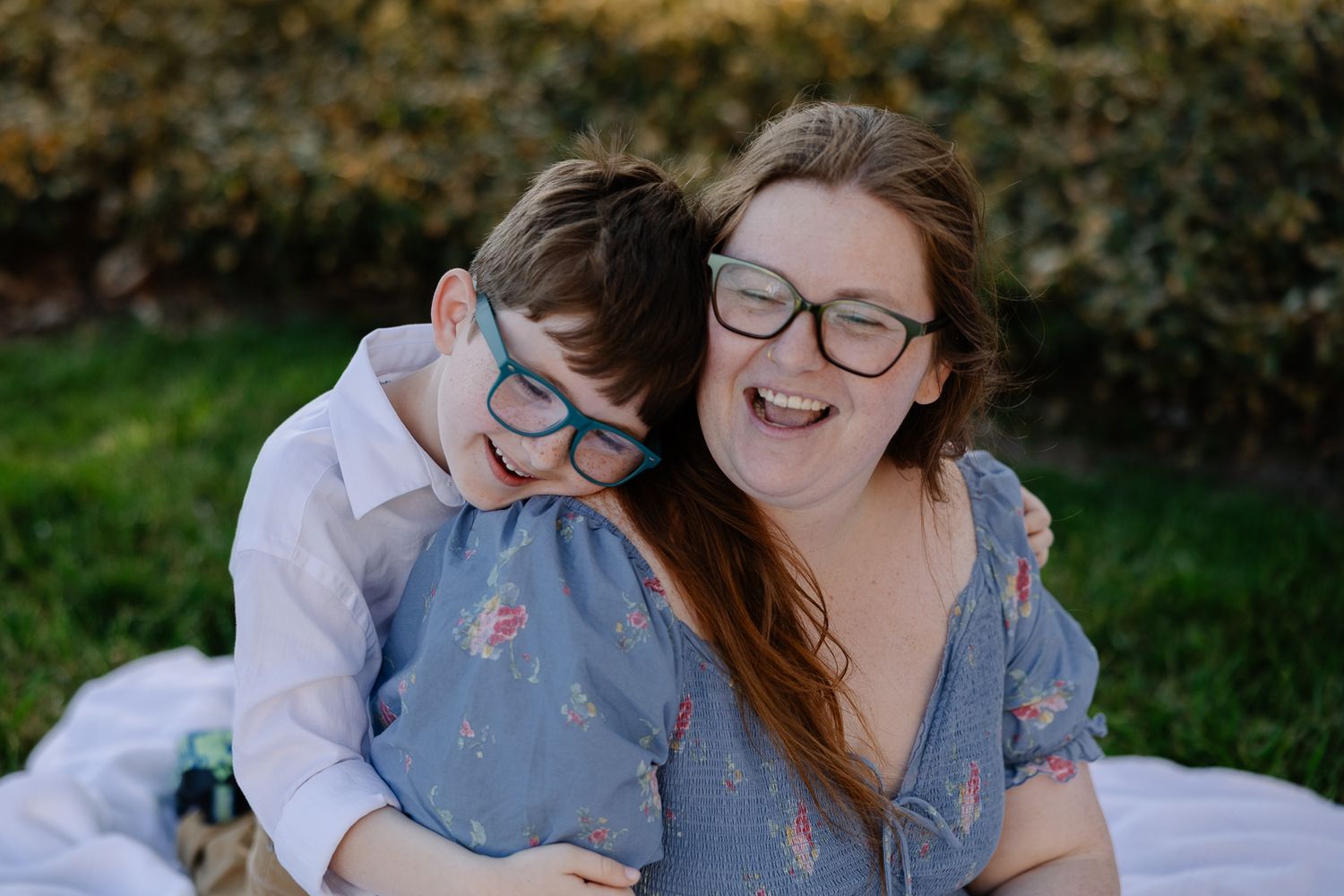 A laughing woman and young boy with matching green glasses share a joyful hug outdoors on a white blanket.