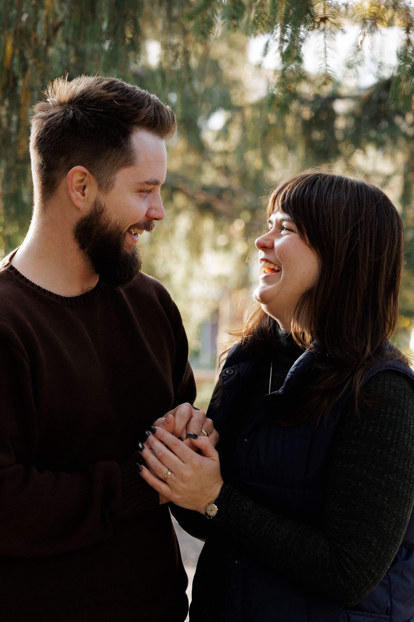 Happy couple holding hands and smiling at each other outdoors near a weeping willow tree.