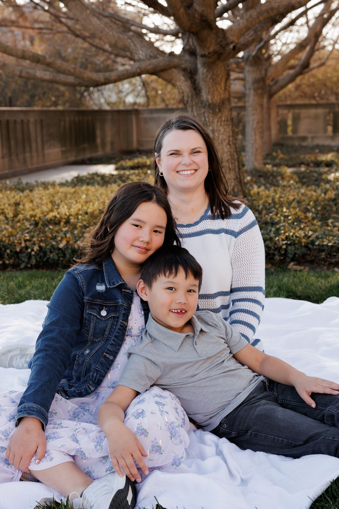A woman and two children smile together on a white blanket outdoors near trees and hedges.