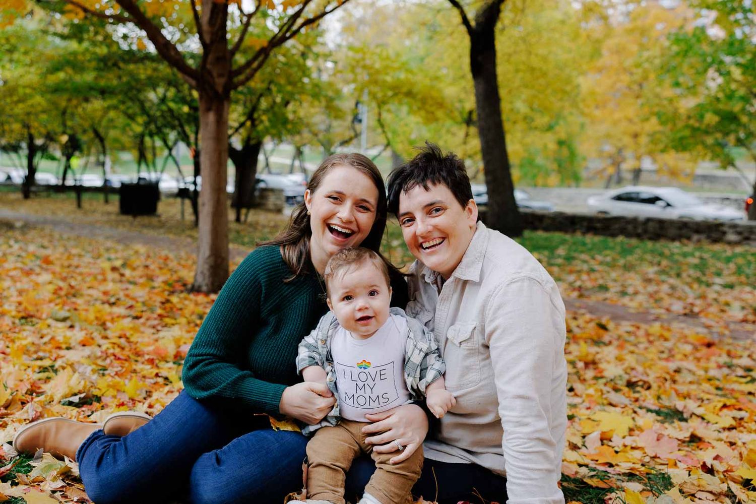 A family smiles together while sitting on colorful fallen leaves during an autumn photoshoot.