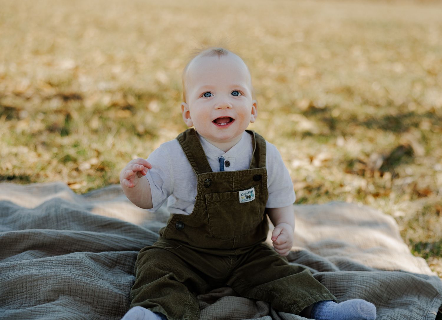 Smiling baby in brown overalls sitting on gray blanket outdoors.