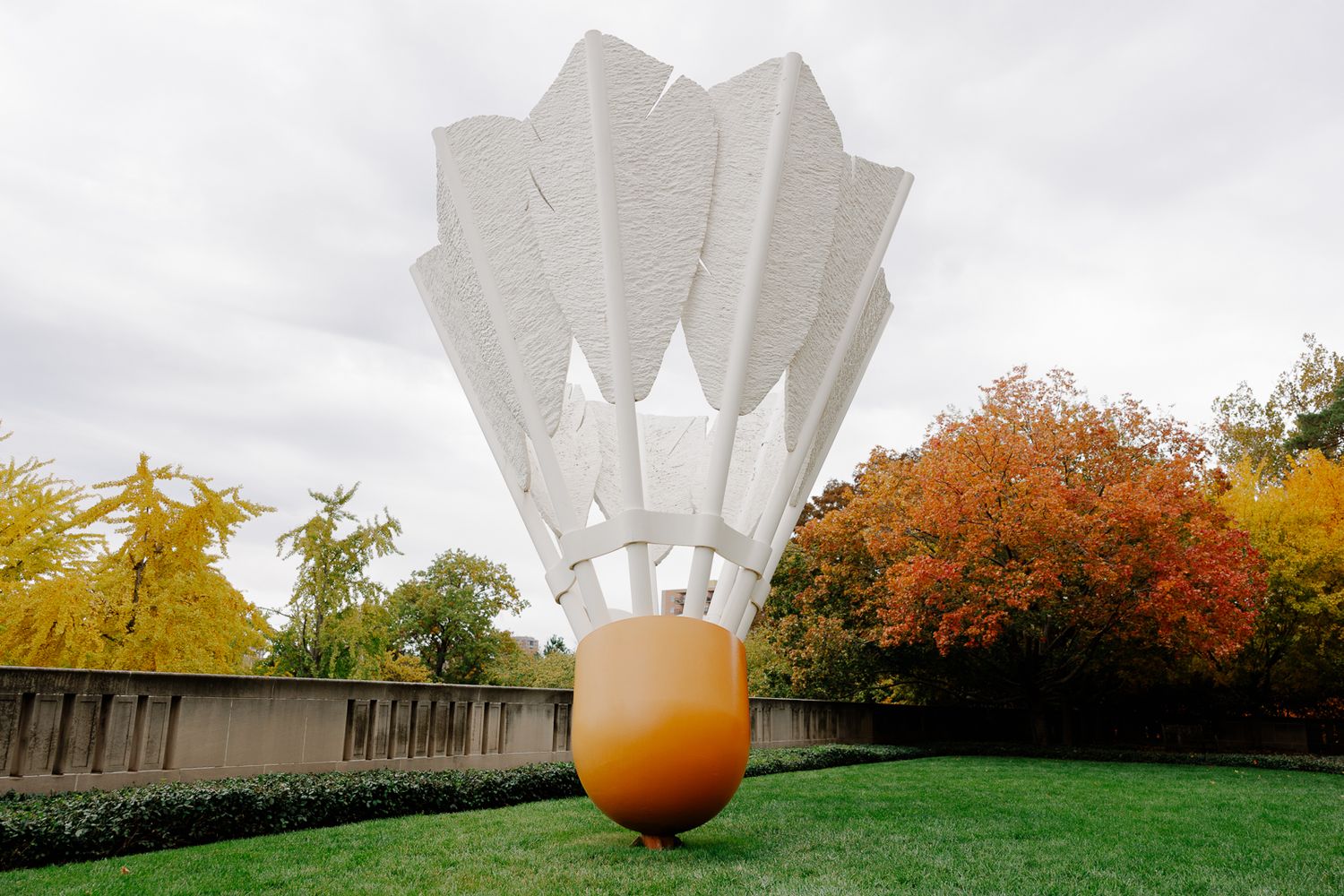 Modern art installation of giant white shuttlecock sculpture against autumn trees.