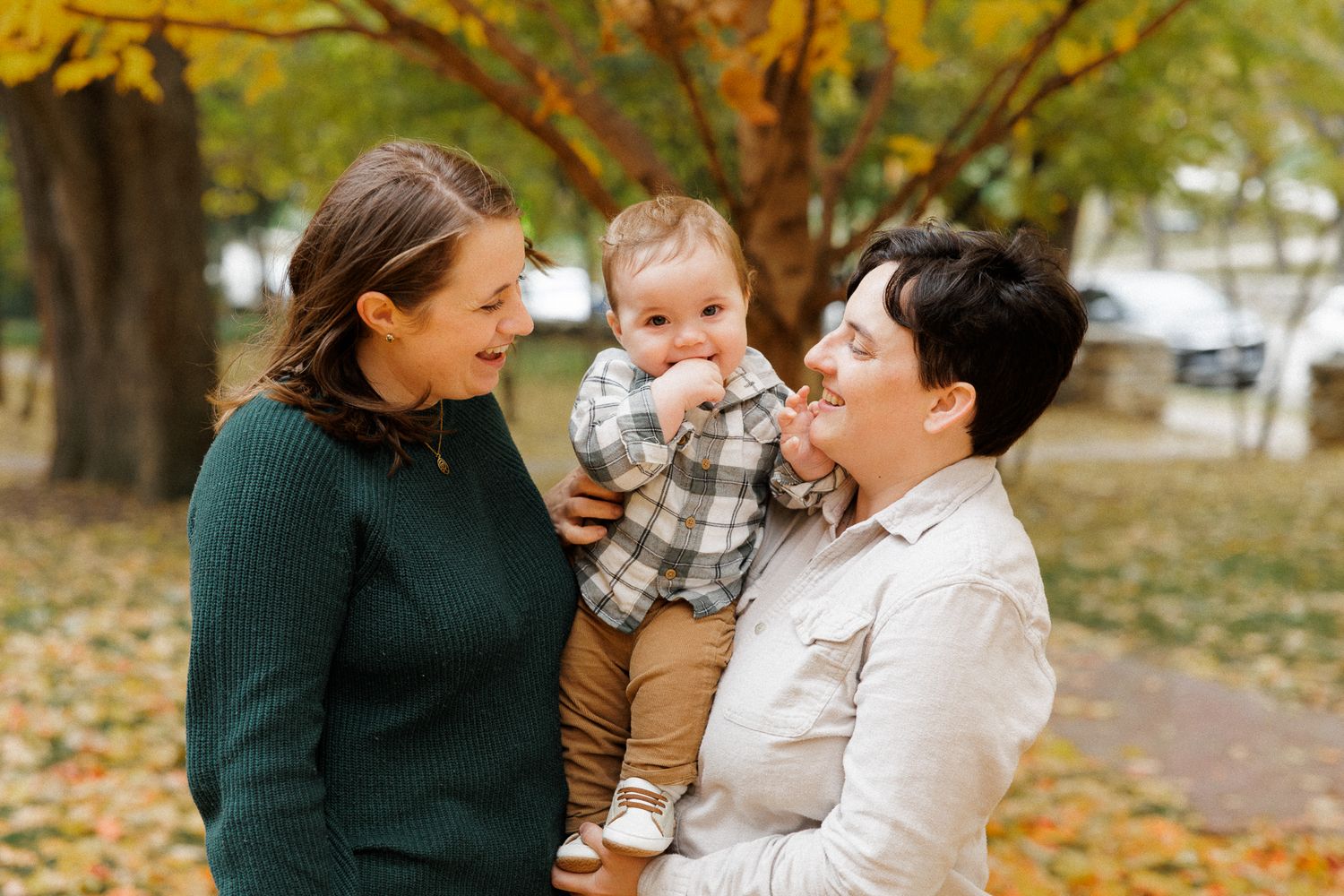 Two women and a baby enjoying an autumn park, smiling together among colorful fallen leaves.