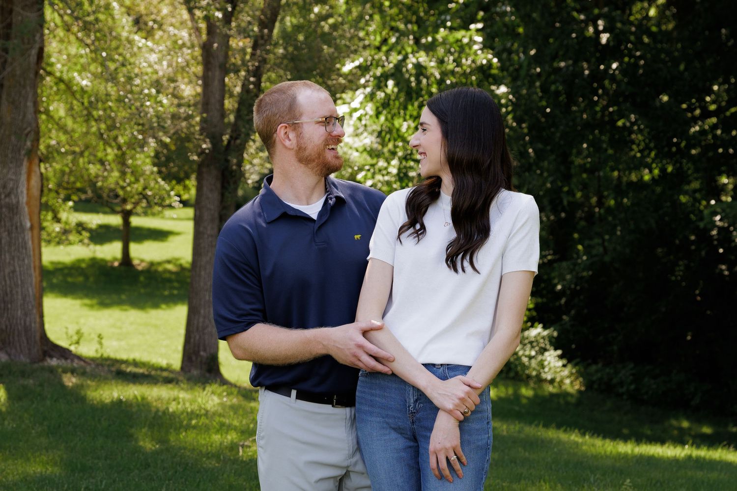 Couple smiling at each other in a sunny green park, man in navy polo and woman in white top and jeans.