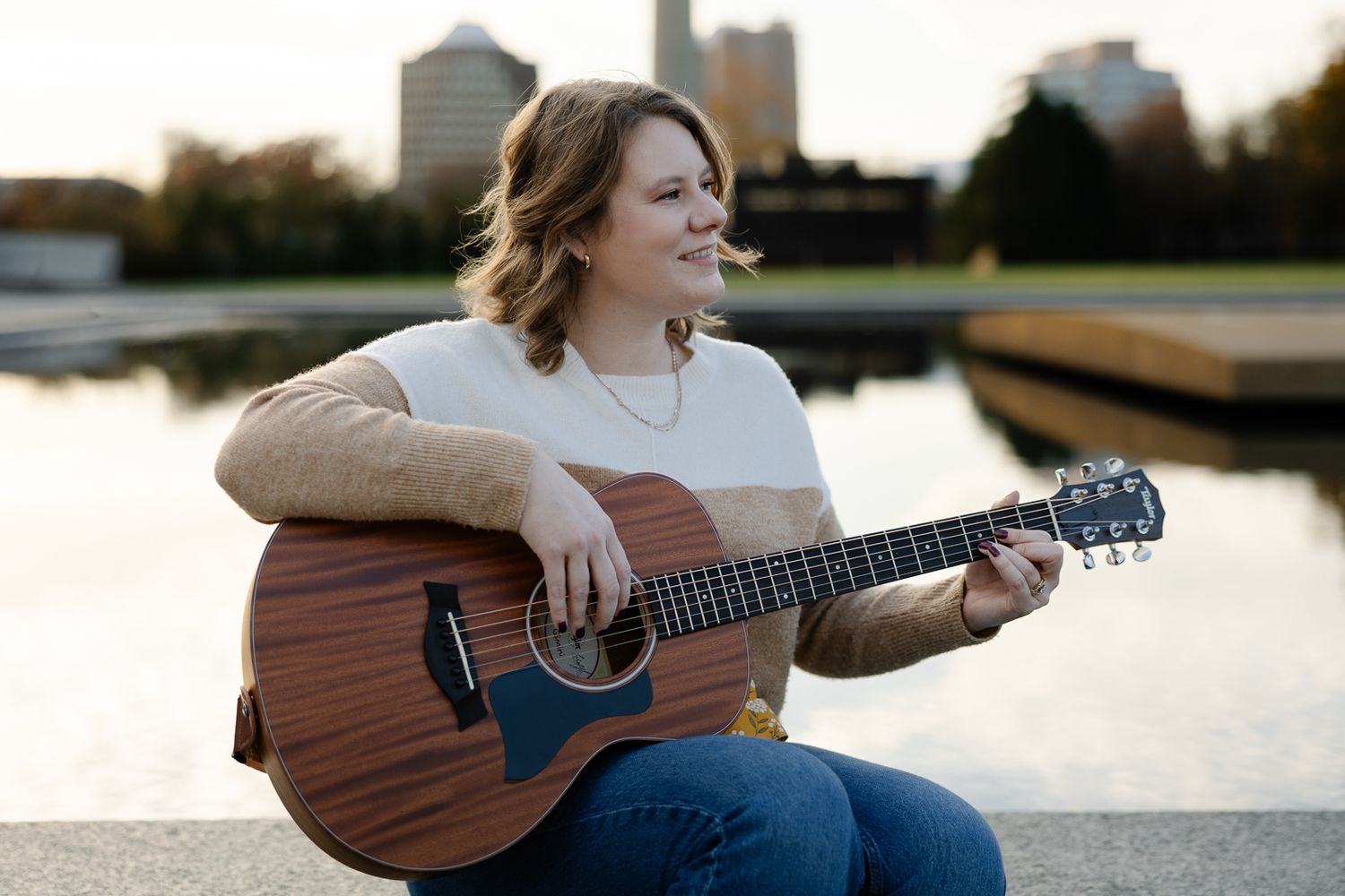 Person sitting by waterfront with guitar during sunset with city skyline in background.