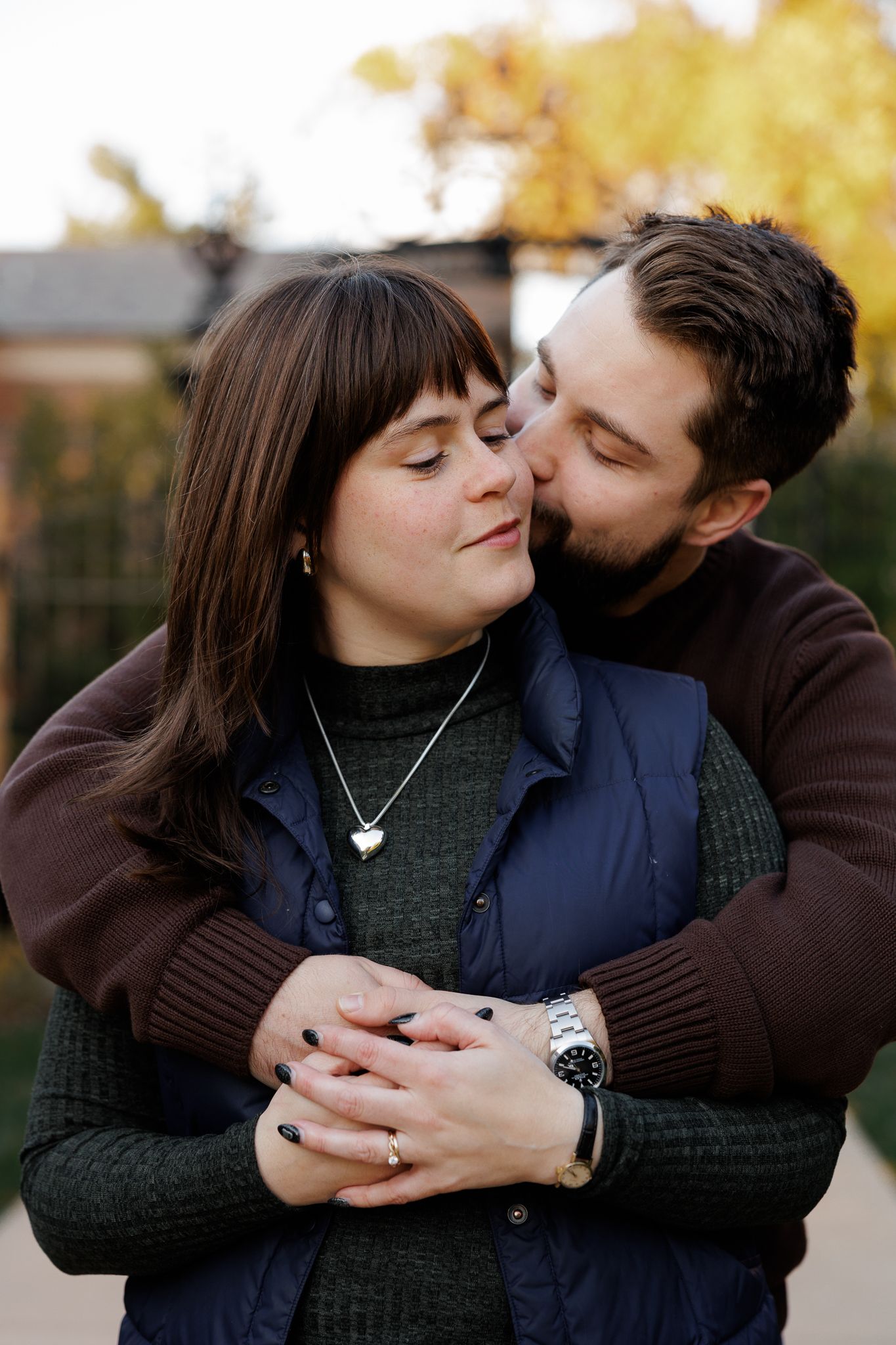 A bearded man embraces a brunette woman from behind, kissing her cheek outdoors in autumn.
