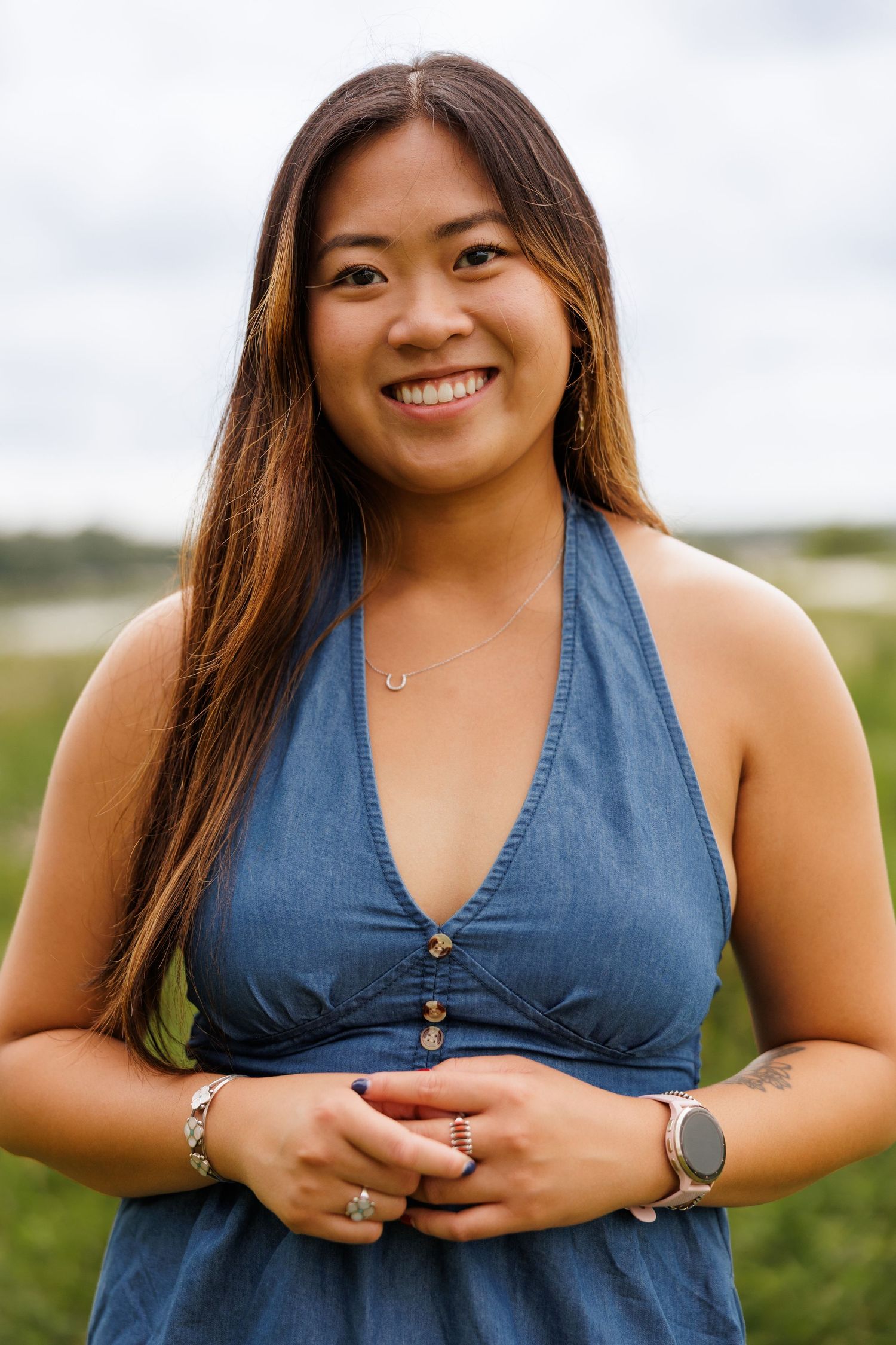Smiling young Asian woman in blue halter dress standing outdoors in a green field on a cloudy day.