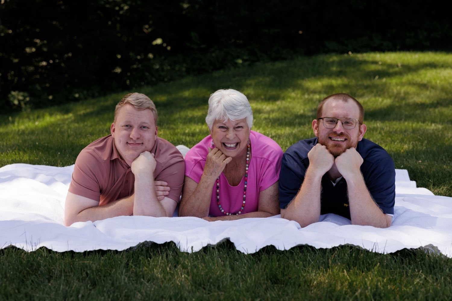 Three people lying on a white blanket on green grass, smiling for a family portrait outdoors.