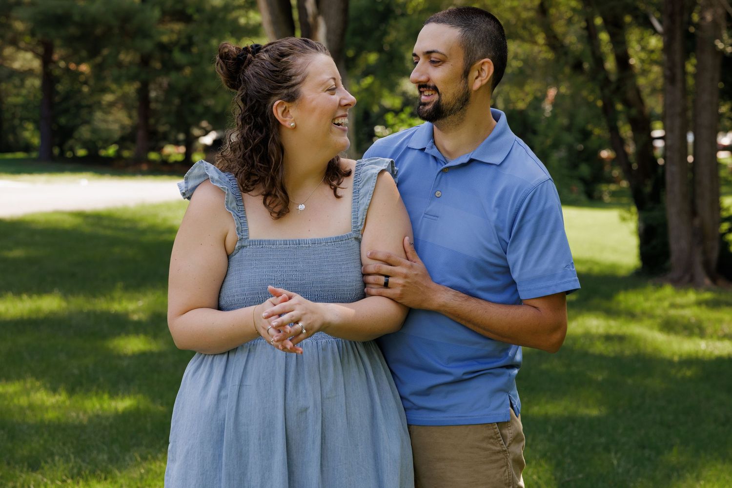 A smiling couple embraces outdoors on a sunny day, woman in blue dress and man in blue polo shirt.