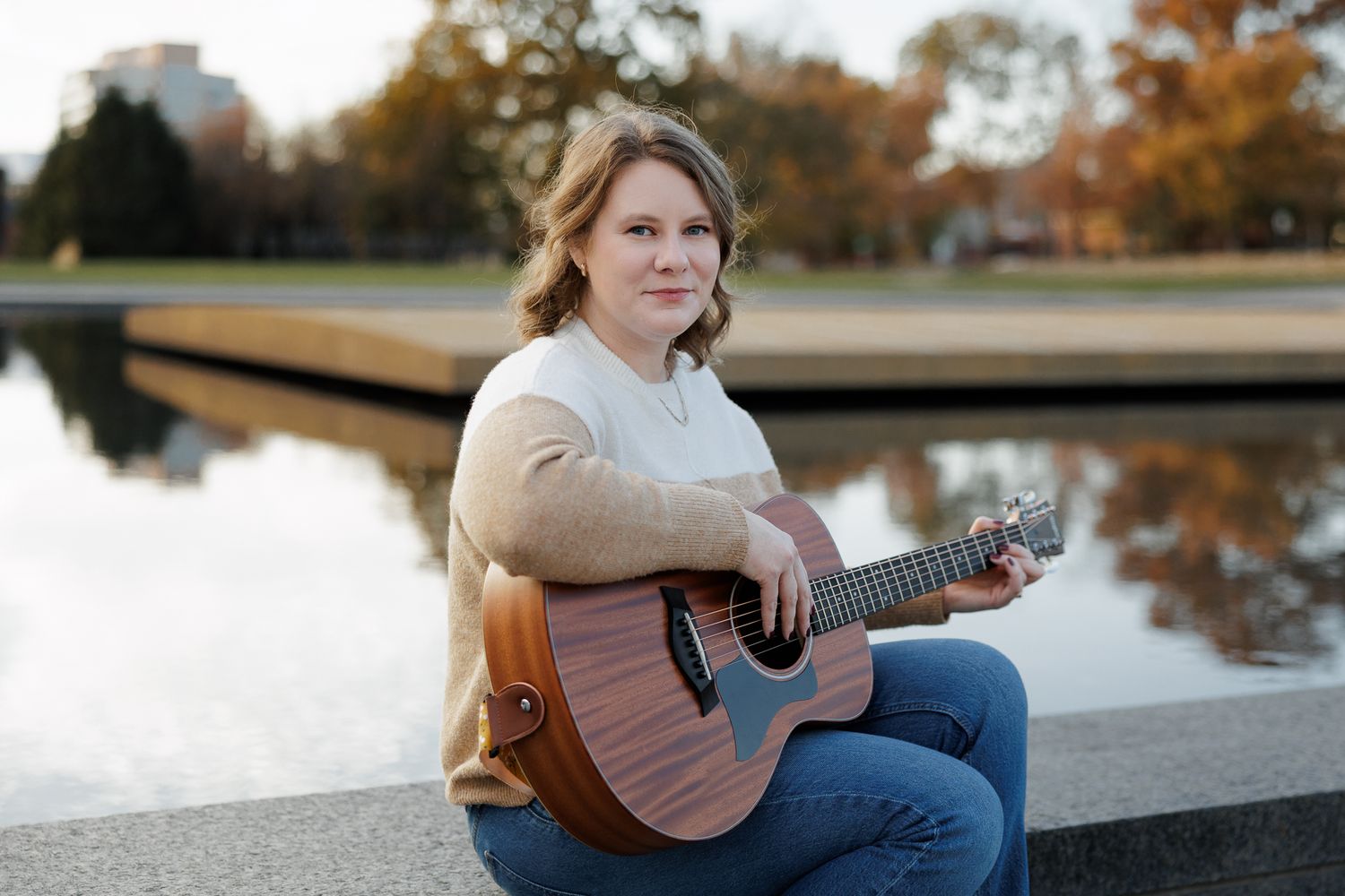 Woman playing acoustic guitar outdoors by a reflective pond in a park during golden hour.