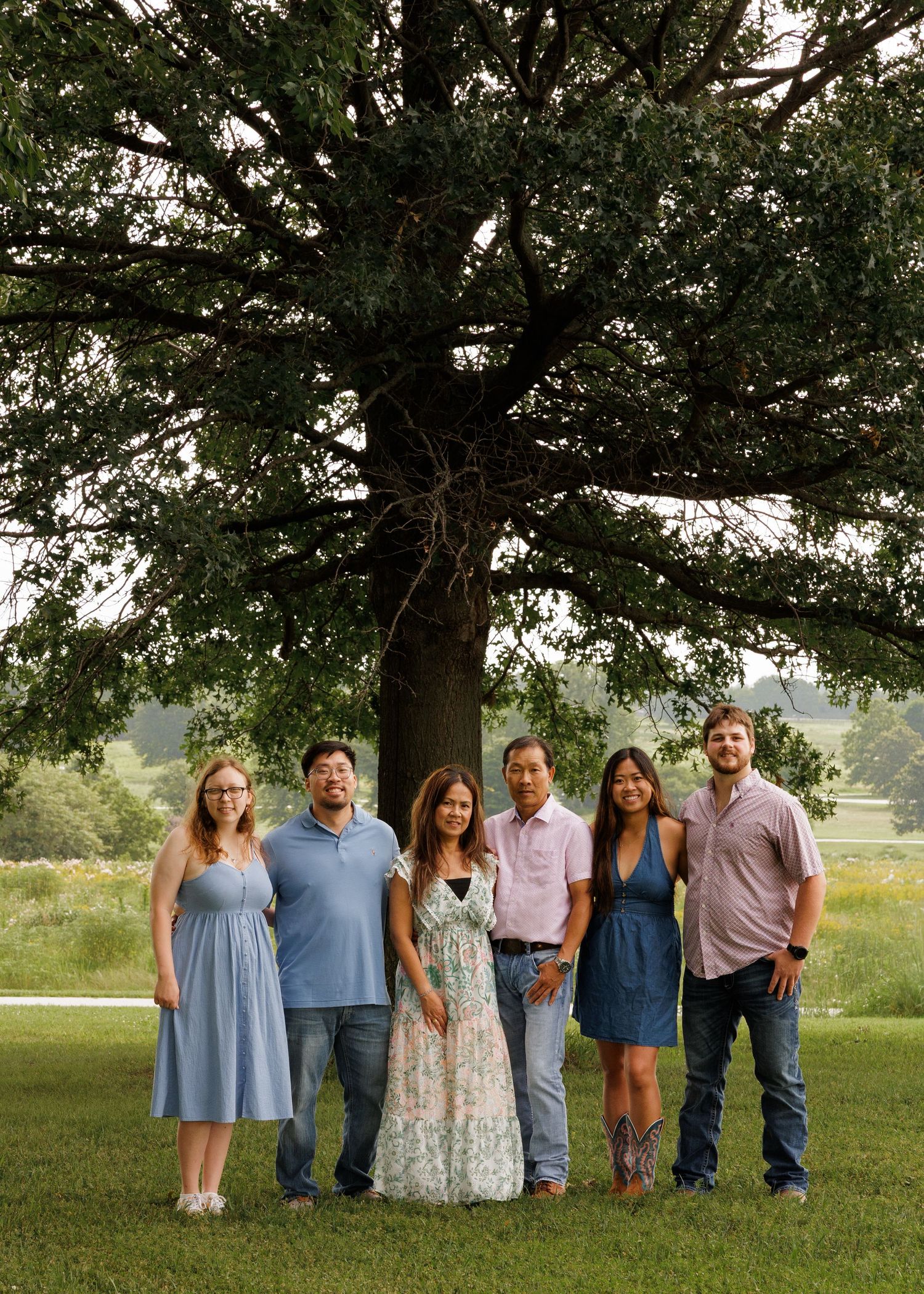 Six people smiling together outdoors under a large oak tree in a green field during a family portrait session.