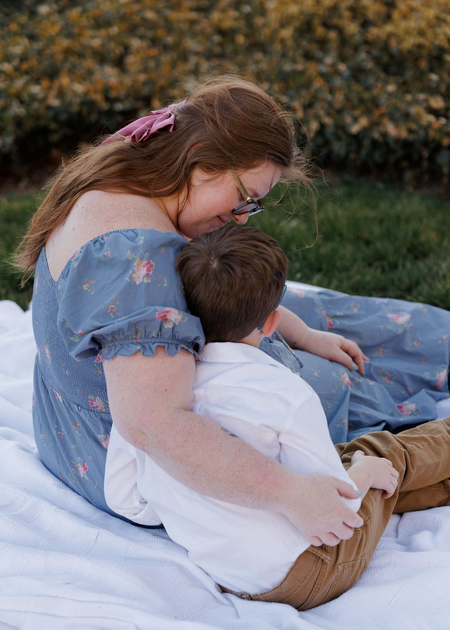 A girl in a floral blue dress hugs a young boy on a white blanket outdoors near green bushes.