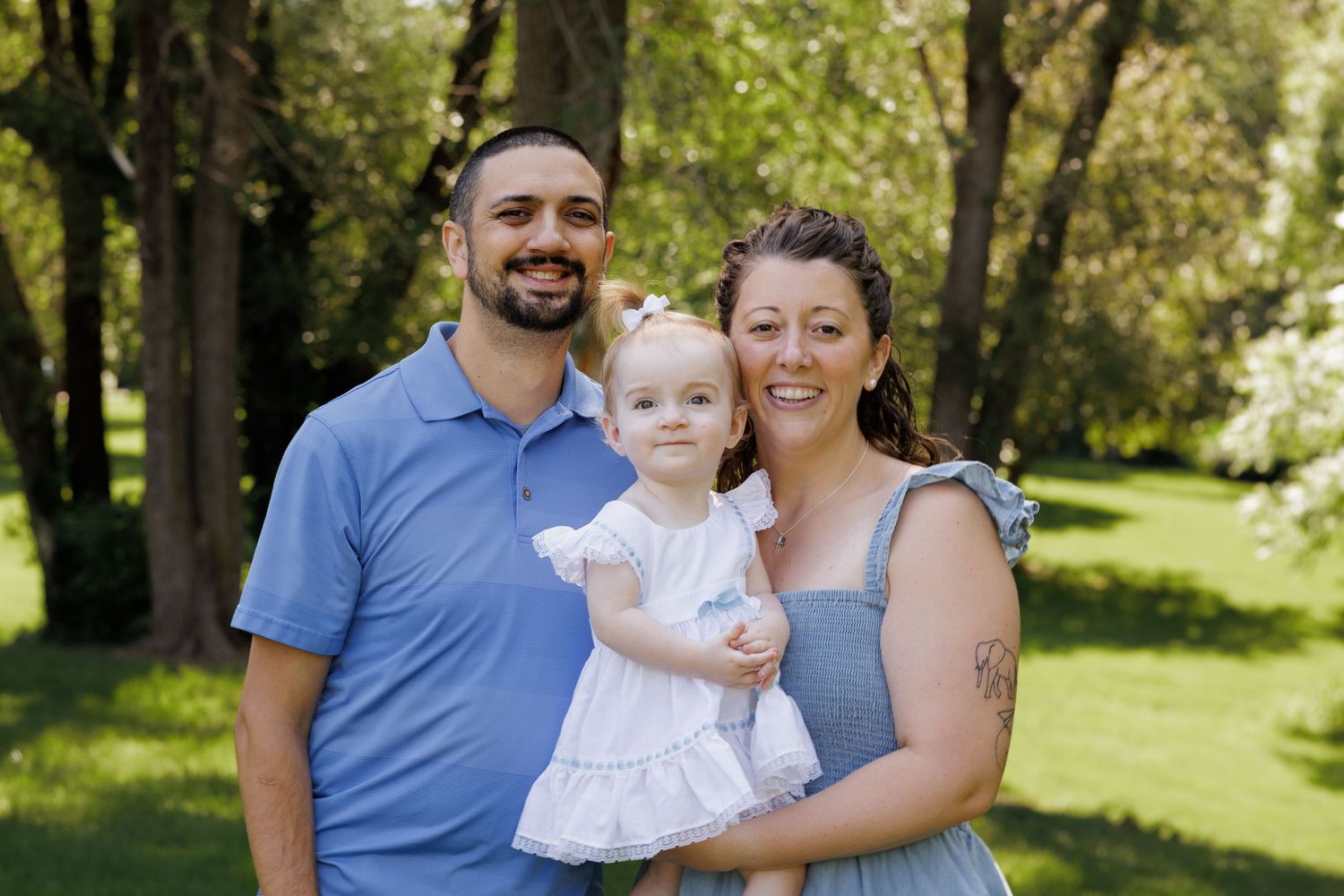 Happy family of three posing outdoors in a lush green park, parents smiling with their toddler daughter in white dress.