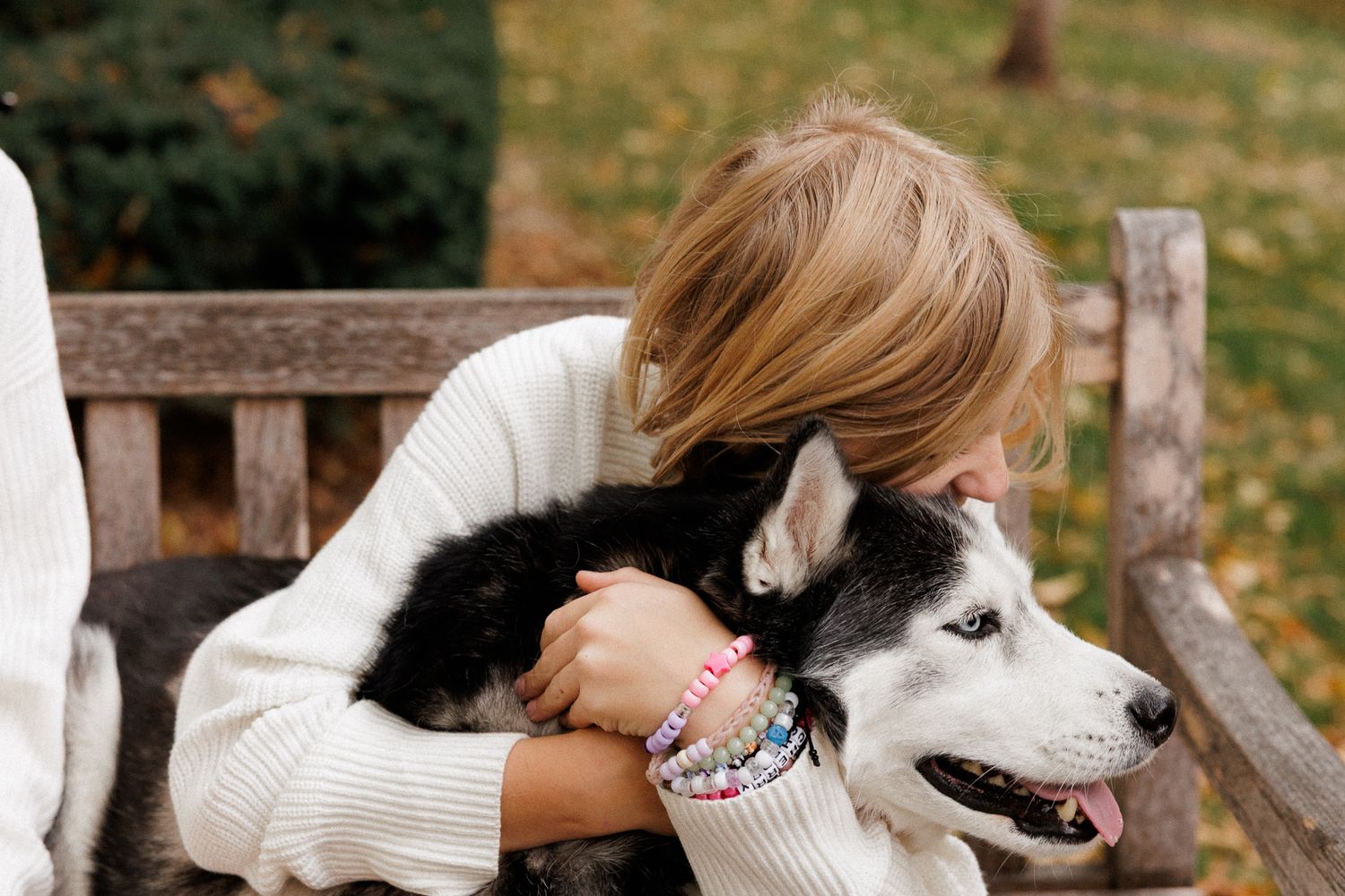 A young blonde girl hugging a black and white Siberian Husky dog on a wooden bench in an autumn park.