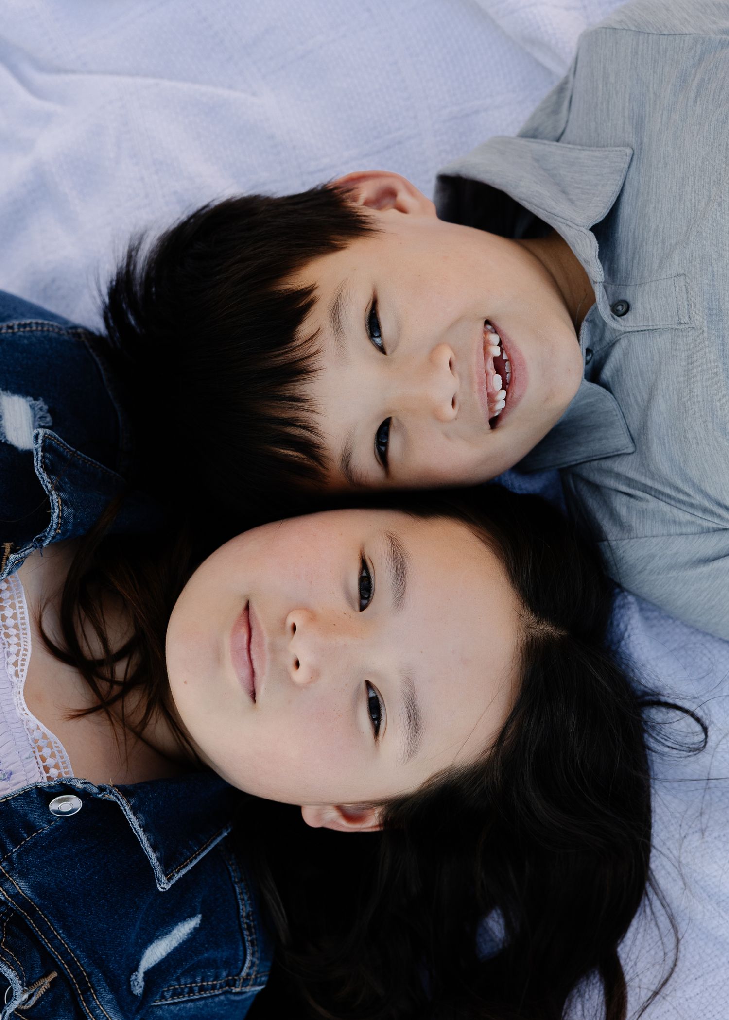 Two smiling children with dark hair lying down together, wearing denim jackets in a candid portrait photo.