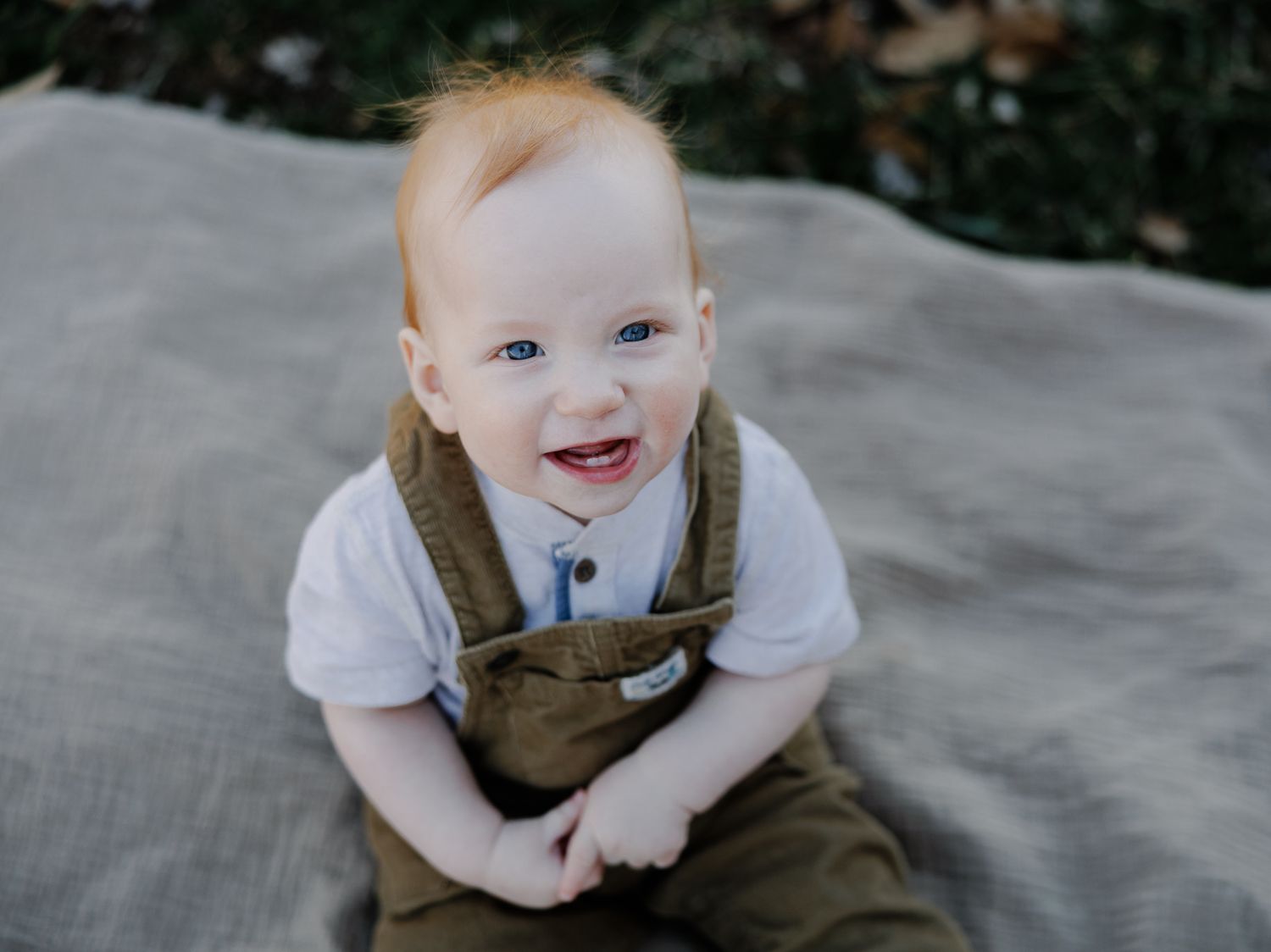 Happy baby wearing brown overalls and white shirt sitting on blanket.
