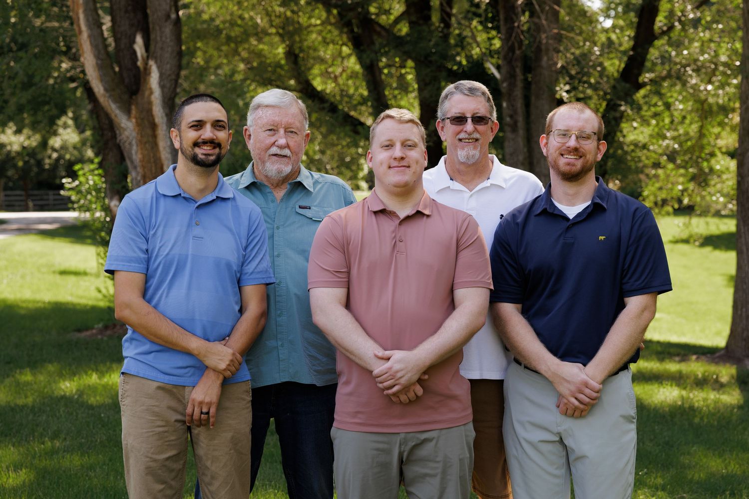 Five men smiling outdoors in a park setting, posing together for a group photo on a sunny day.