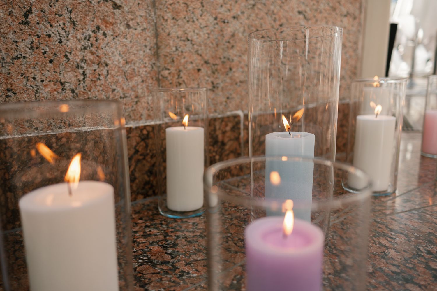 Lit pillar candles in white, gray, and purple displayed on a granite surface with glass hurricane holders.