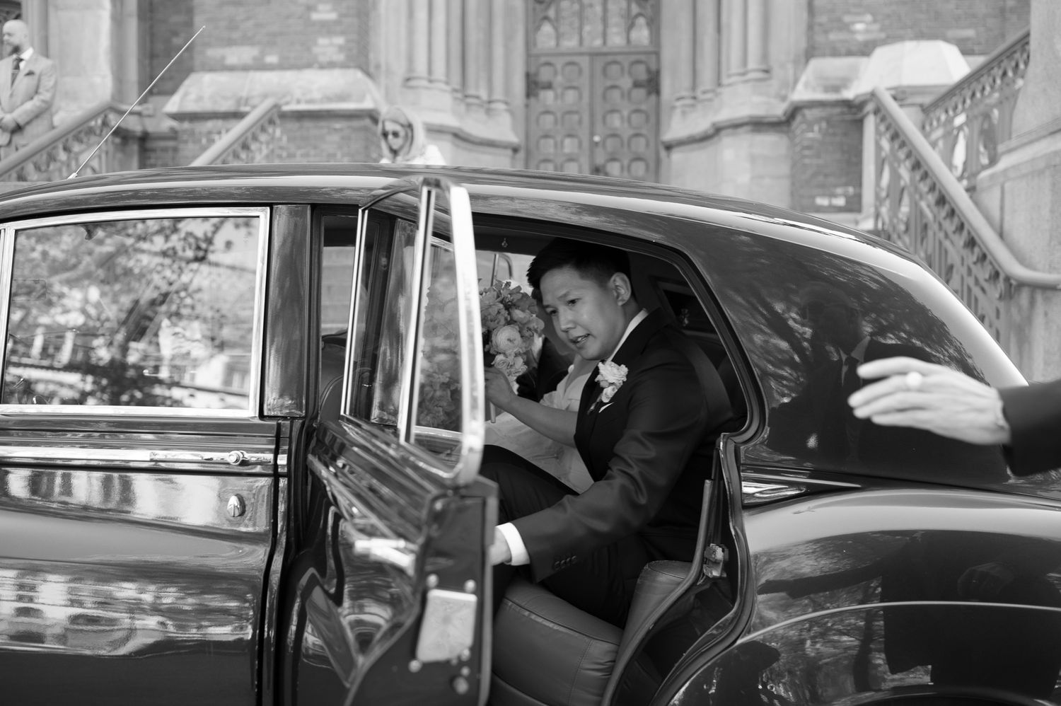 Groom in tuxedo stepping out of a classic black wedding car outside a church in black and white photo.