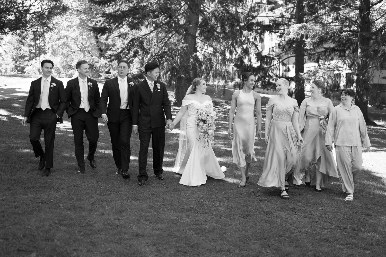 Black and white photo of a wedding party walking outdoors, with groomsmen in tuxedos and bridesmaids in elegant gowns.