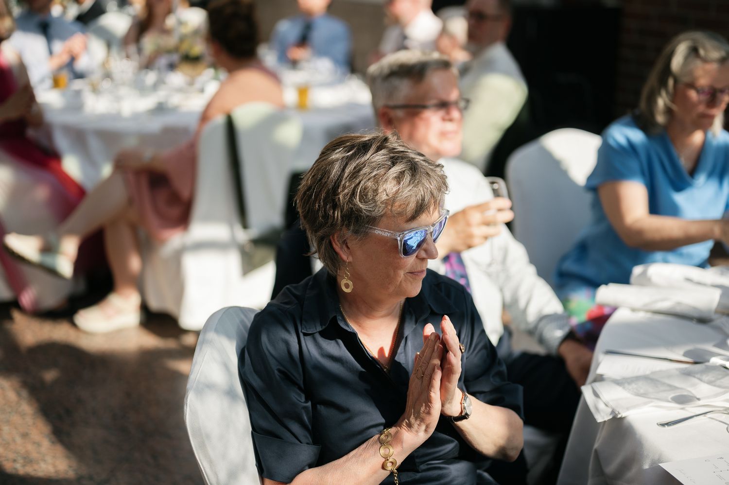 Woman in sunglasses with hands in prayer gesture at an outdoor wedding reception on a sunny day.