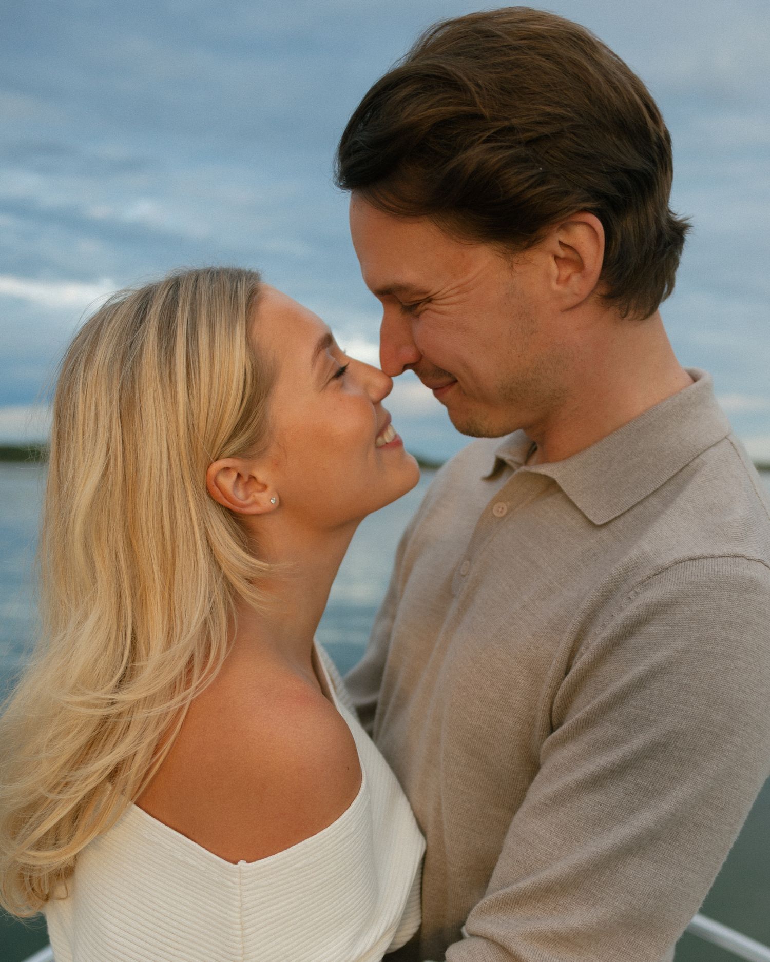 A romantic couple sharing an intimate moment, nearly kissing outdoors near water with a cloudy sky backdrop.