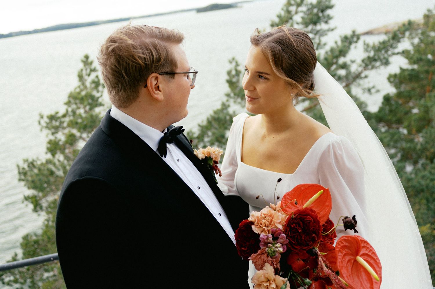Bride and groom share intimate moments during outdoor wedding photoshoot with orange and red floral bouquet.