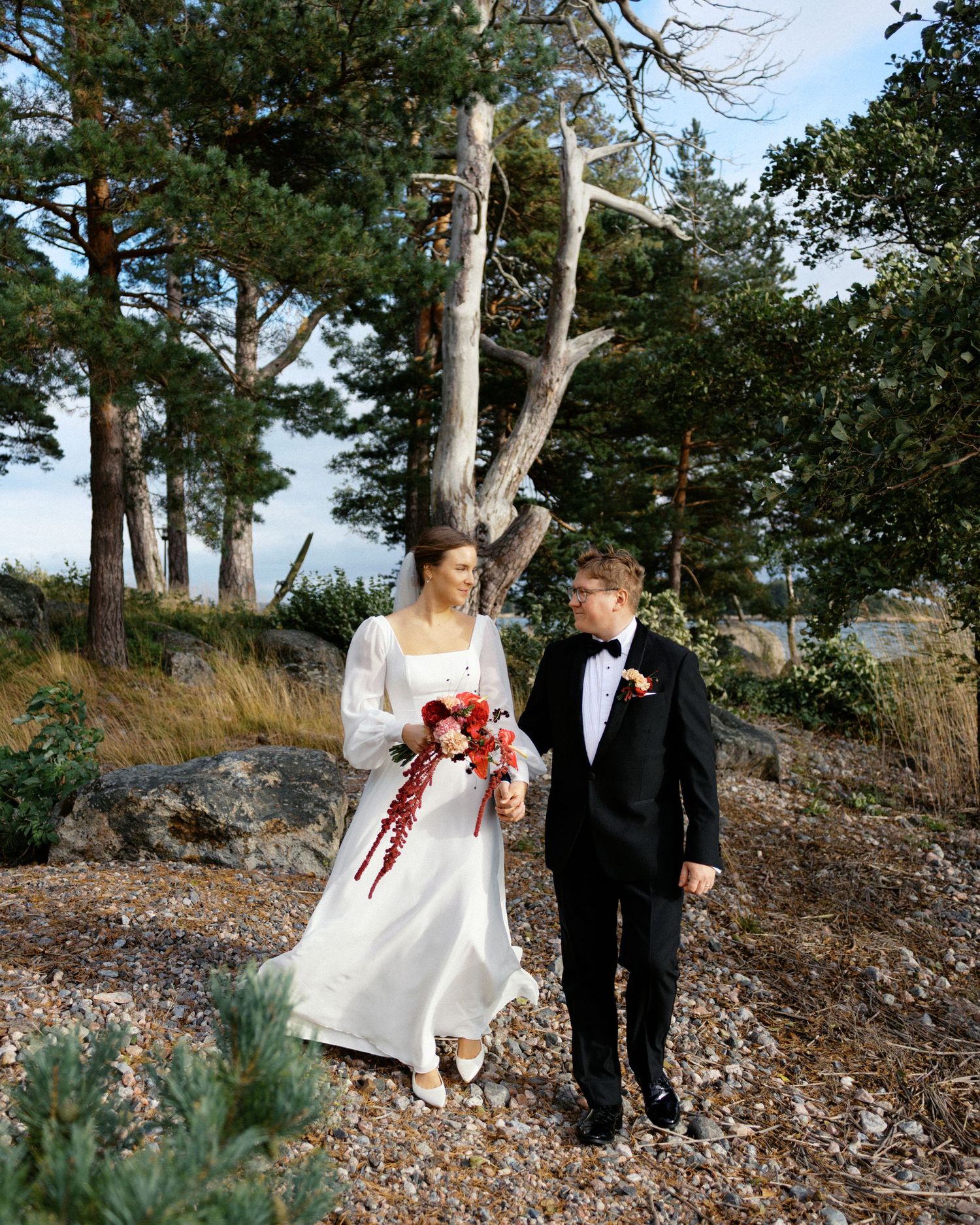 A couple in wedding attire walking together on a rocky coastal path surrounded by pine trees.