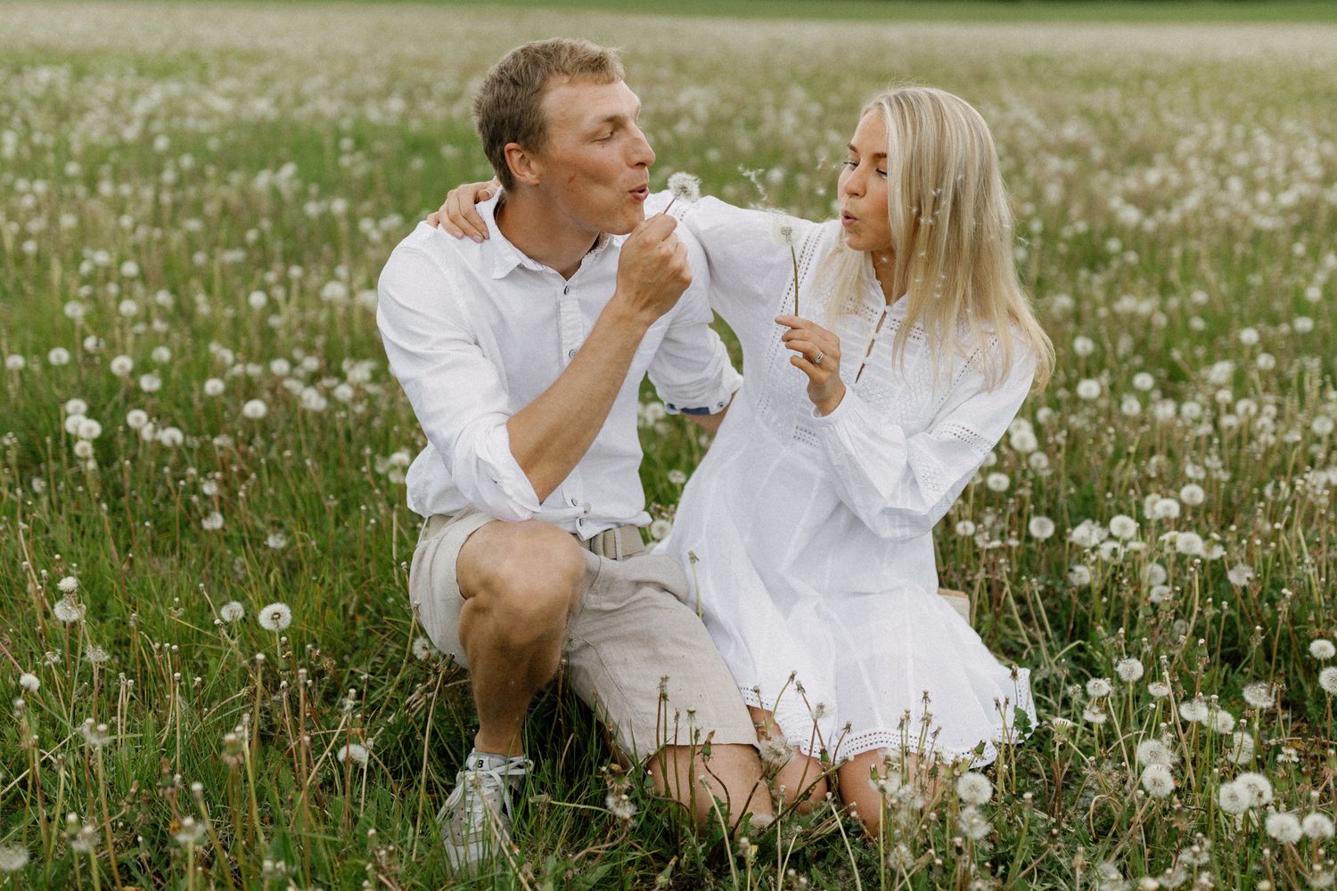 A couple shares a playful moment sitting in a field of white wildflowers.