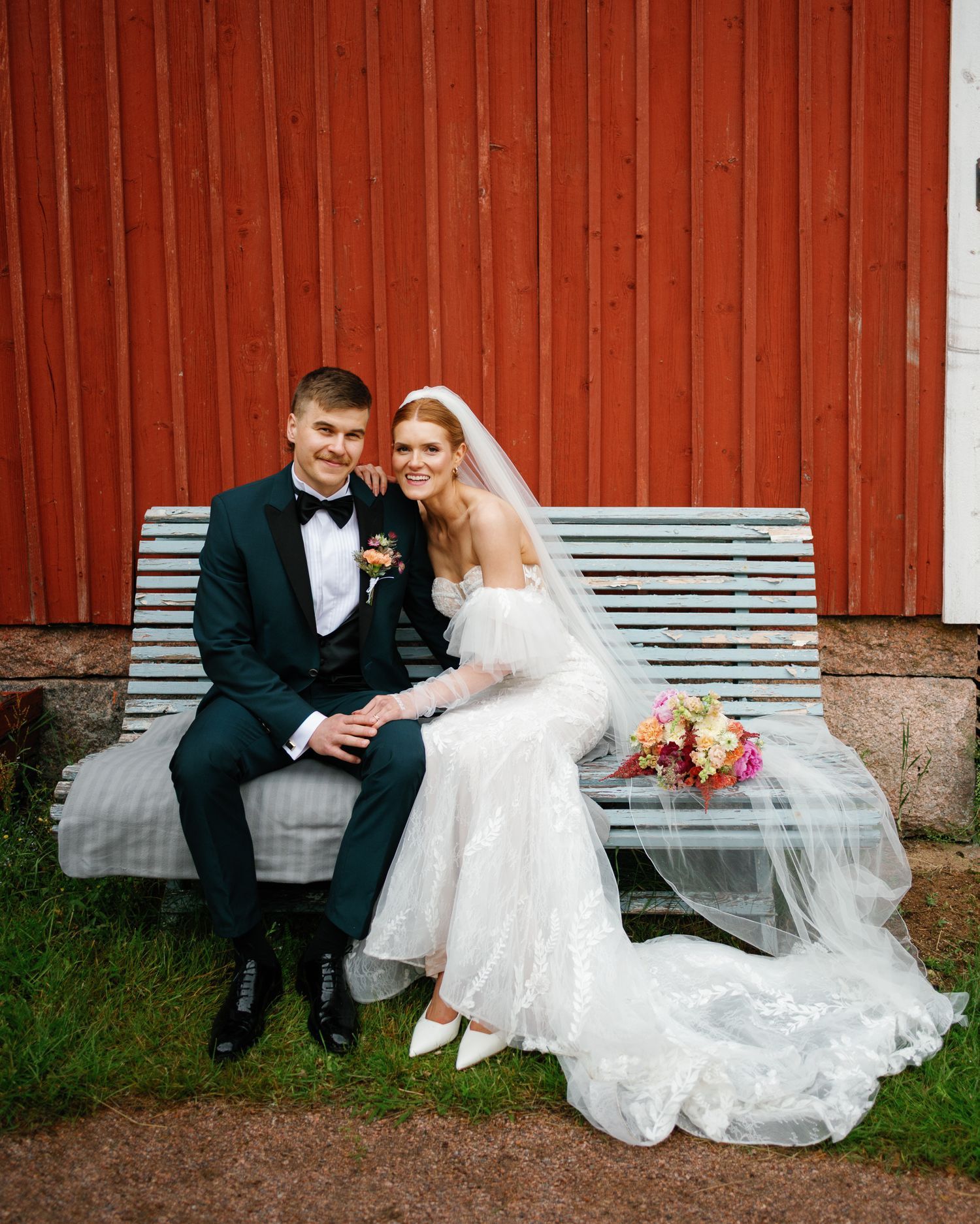 Wedding couple sitting on bench by red barn wall with flowing white wedding dress.