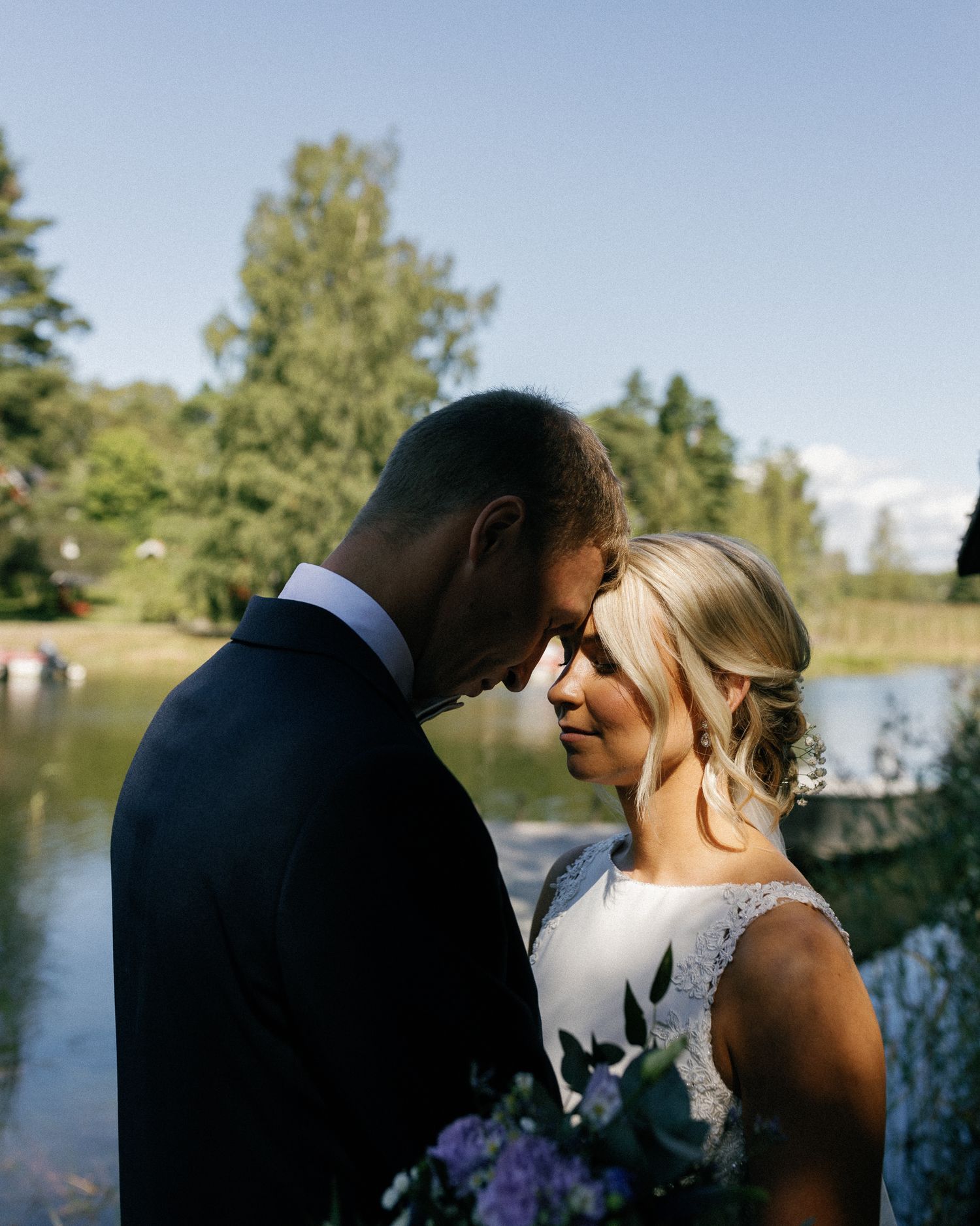 Wedding couple shares tender moments by a serene lake with trees in the background.