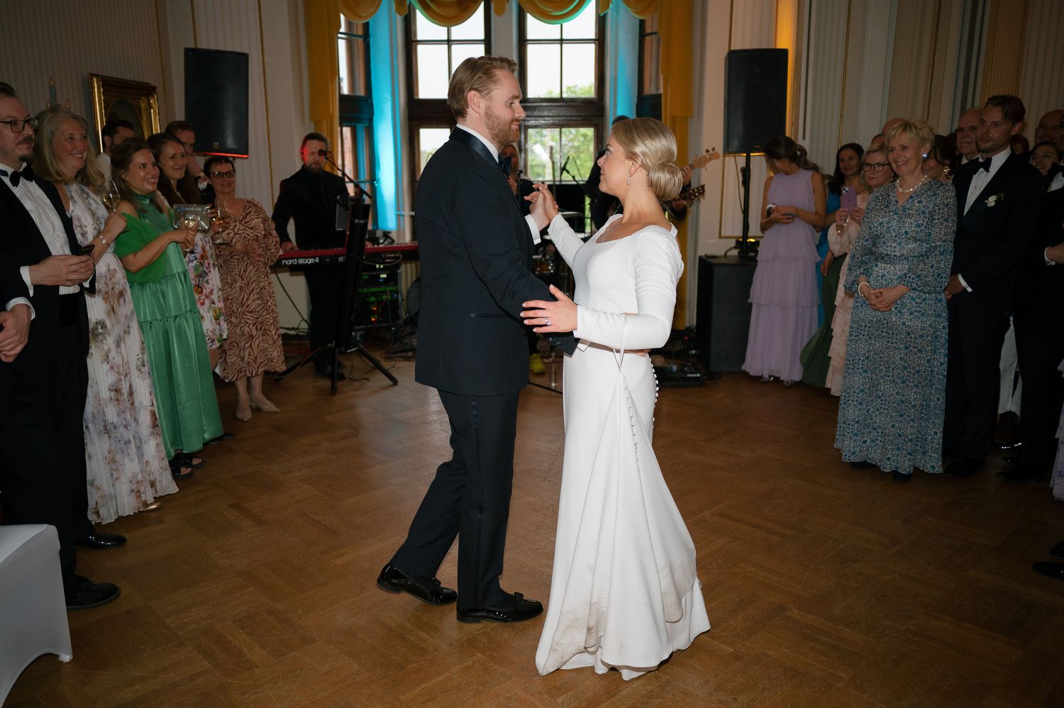 Couples dancing together at an elegant reception in a grand ballroom with large windows and wooden floors.
