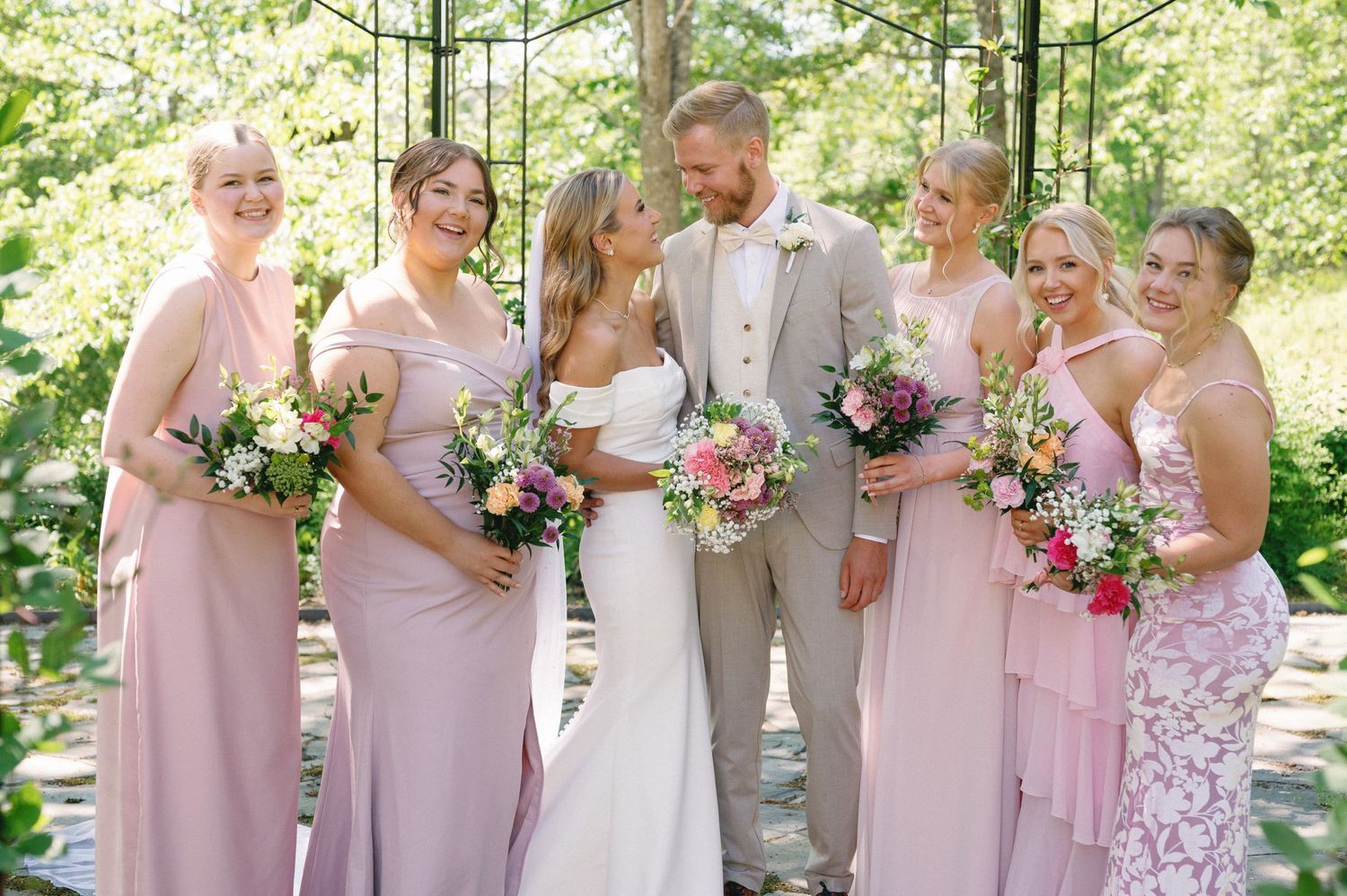 Bridal party in pink dresses holds floral bouquets in outdoor garden setting.
