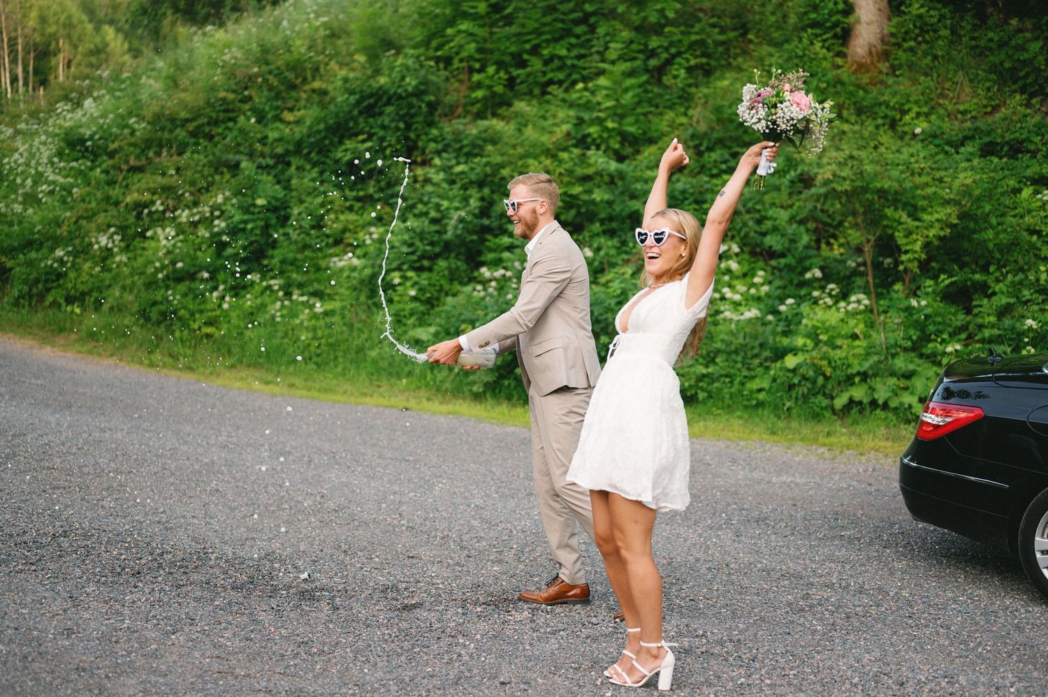 People celebrate on a gravel path surrounded by green foliage and parked cars.