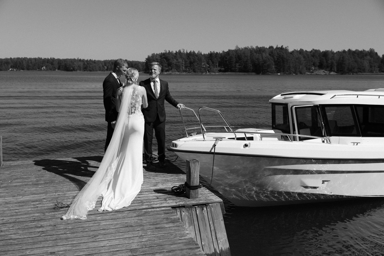 A couple stands on a wooden dock next to a white boat on a serene lake.