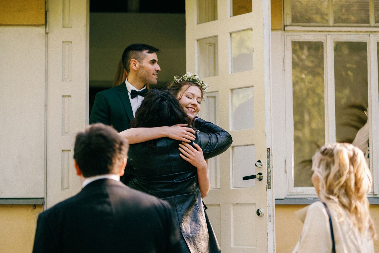 Wedding guests gather outside a building with white doors and yellow walls.