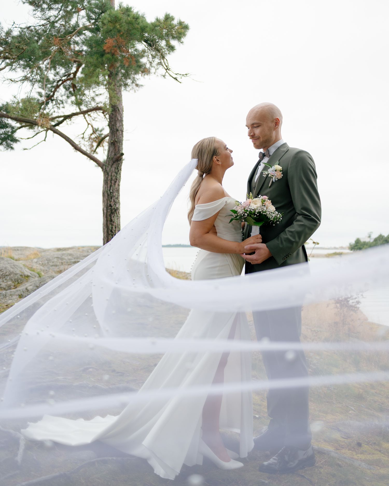 Couple in wedding attire posing on rocky terrain with flowing veil catching the wind.