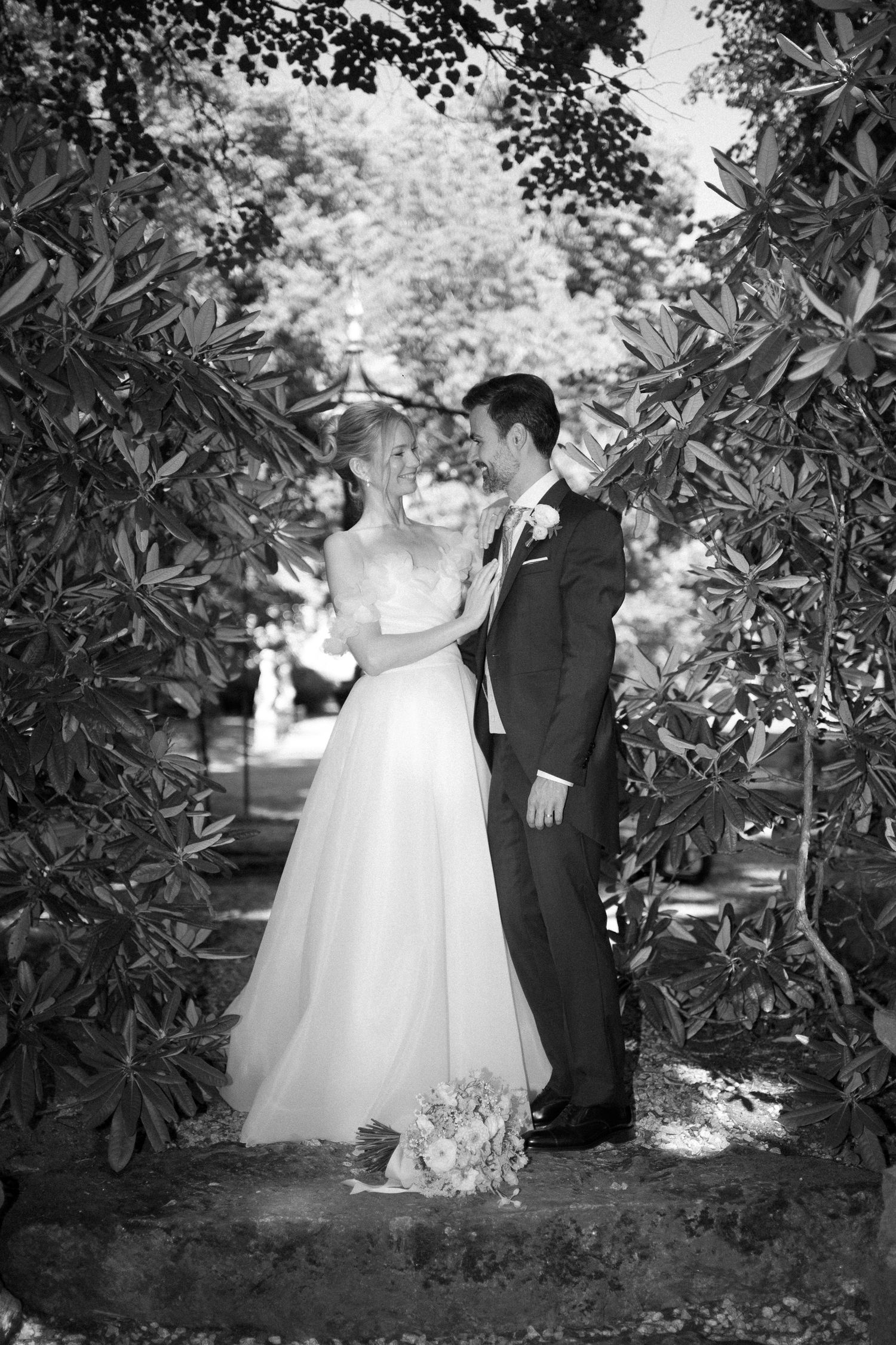 Black and white wedding photo of bride and groom sharing romantic moment framed by lush garden archway.