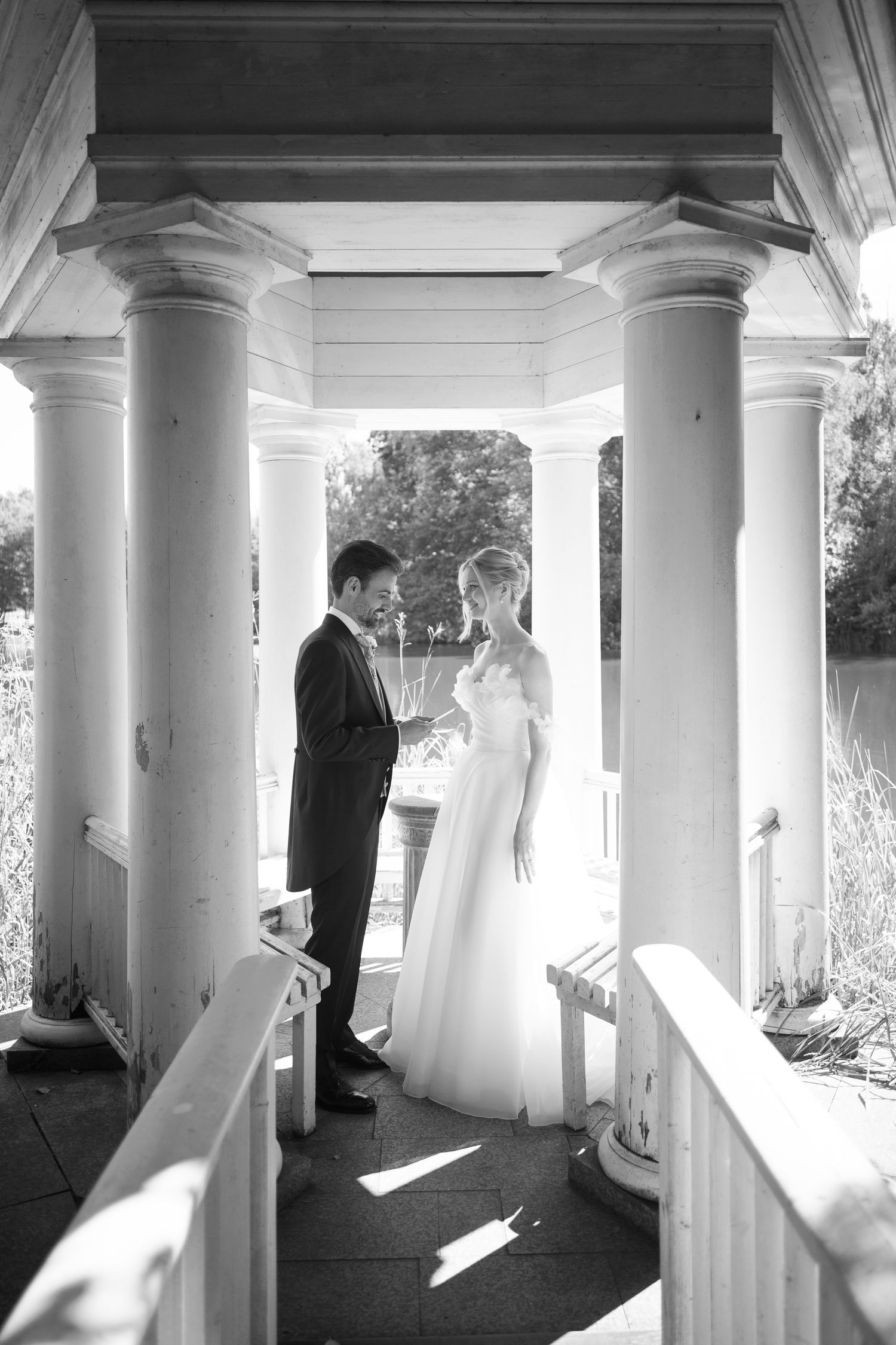 Black and white photo of bride and groom standing between elegant columns in a classical gazebo on their wedding day.