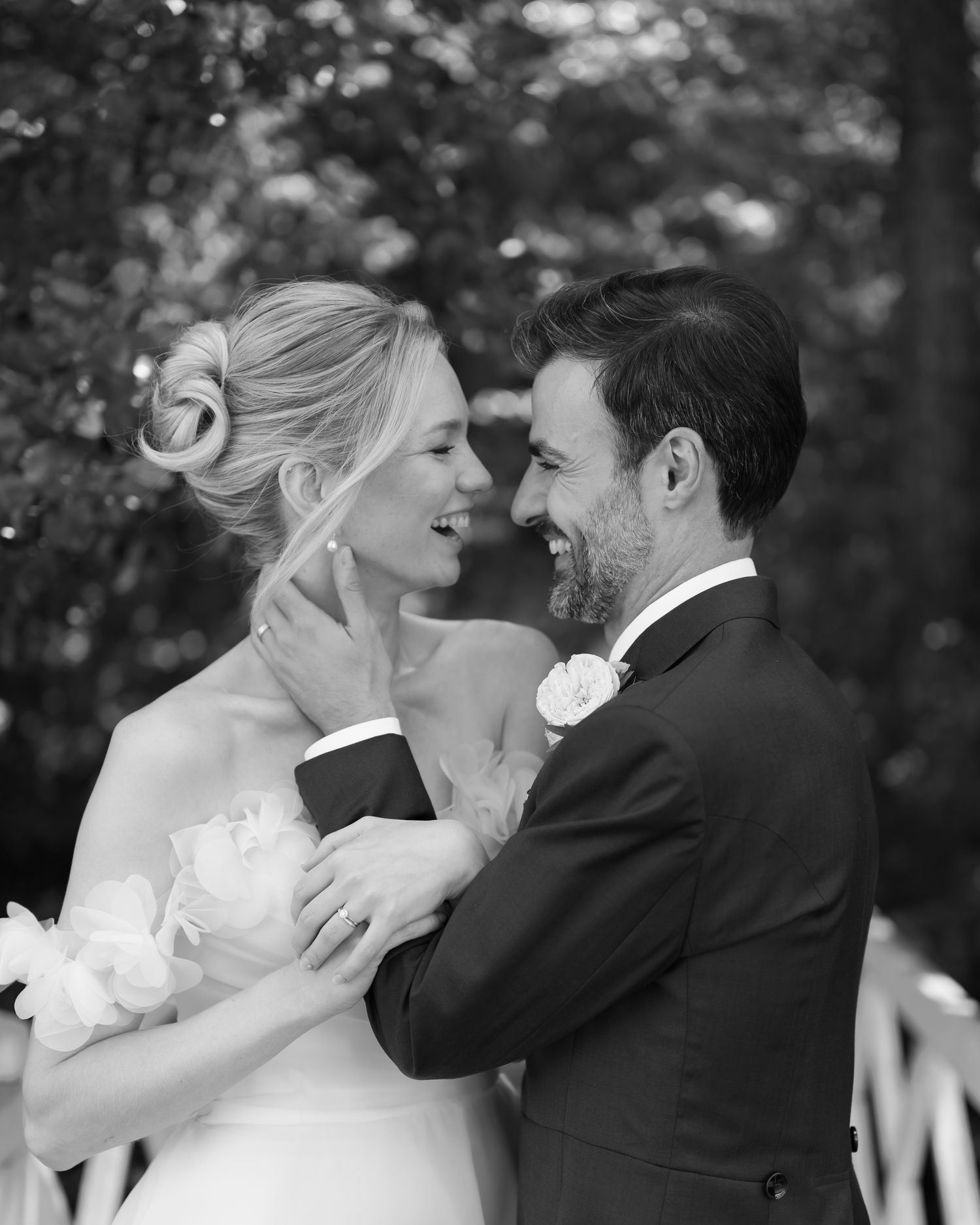 Black and white wedding photo of smiling bride and groom sharing a tender moment outdoors amid lush trees.