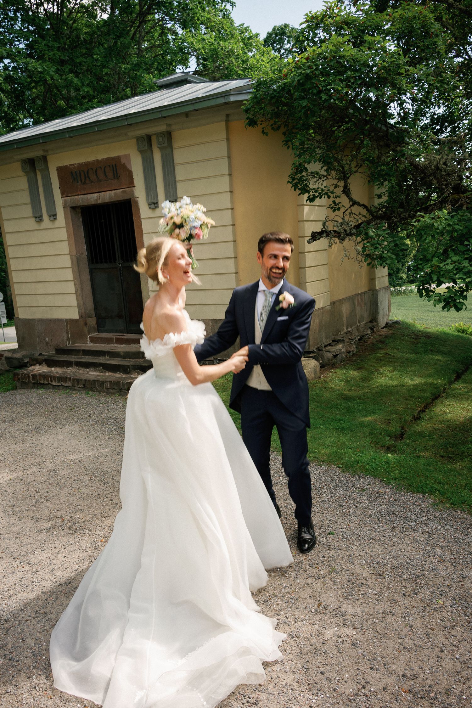 A joyful bride in a white gown and groom in navy suit hold hands walking outside a yellow building.