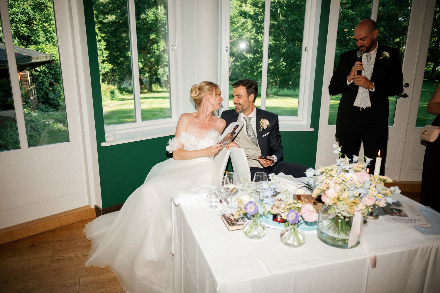 Bride and groom seated at wedding reception table, smiling and reading speeches, with floral centerpiece and guest in background.
