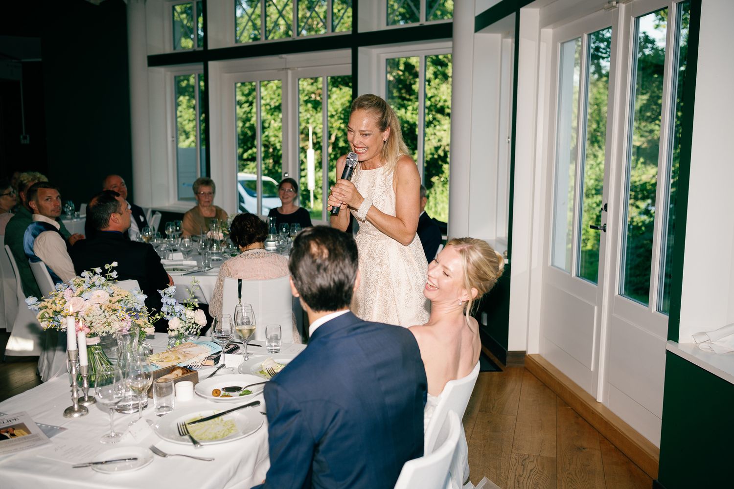Woman giving a speech at an elegant wedding reception dinner in a bright, green-windowed venue.