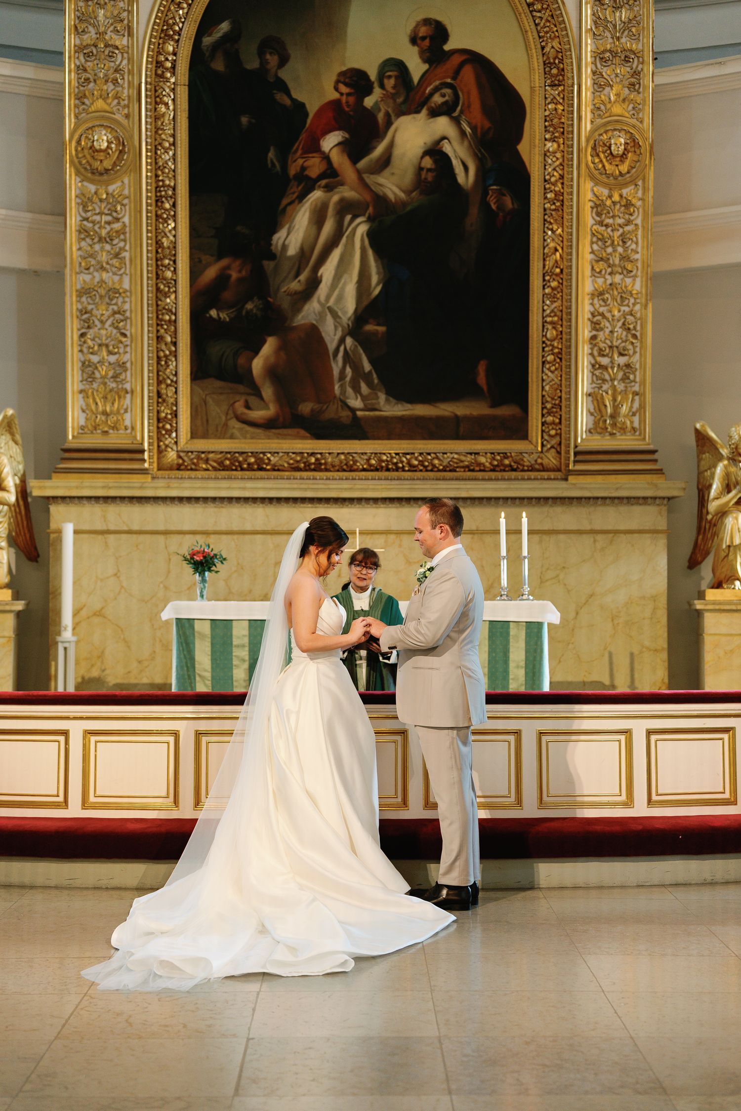 Bride and groom exchange rings at church altar during wedding ceremony with large religious painting backdrop.